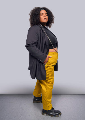 1X=A person with curly hair confidently poses, wearing a Debbie Harry X Wildfang Double Breasted Blazer over a black top and bright yellow pants. The outfit, featuring subtle metal detailing, complements their stance on a grey floor against a plain white backdrop.