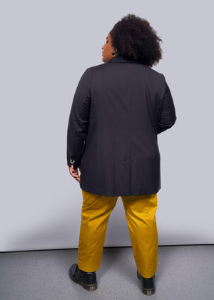 1X= Showcasing a Debbie Harry X Wildfang Double Breasted Blazer with subtle metal details, the person pairs it stylishly with mustard yellow pants and black shoes. Their curly hair cascades down their back as they stand against a pristine white background on a light gray floor.