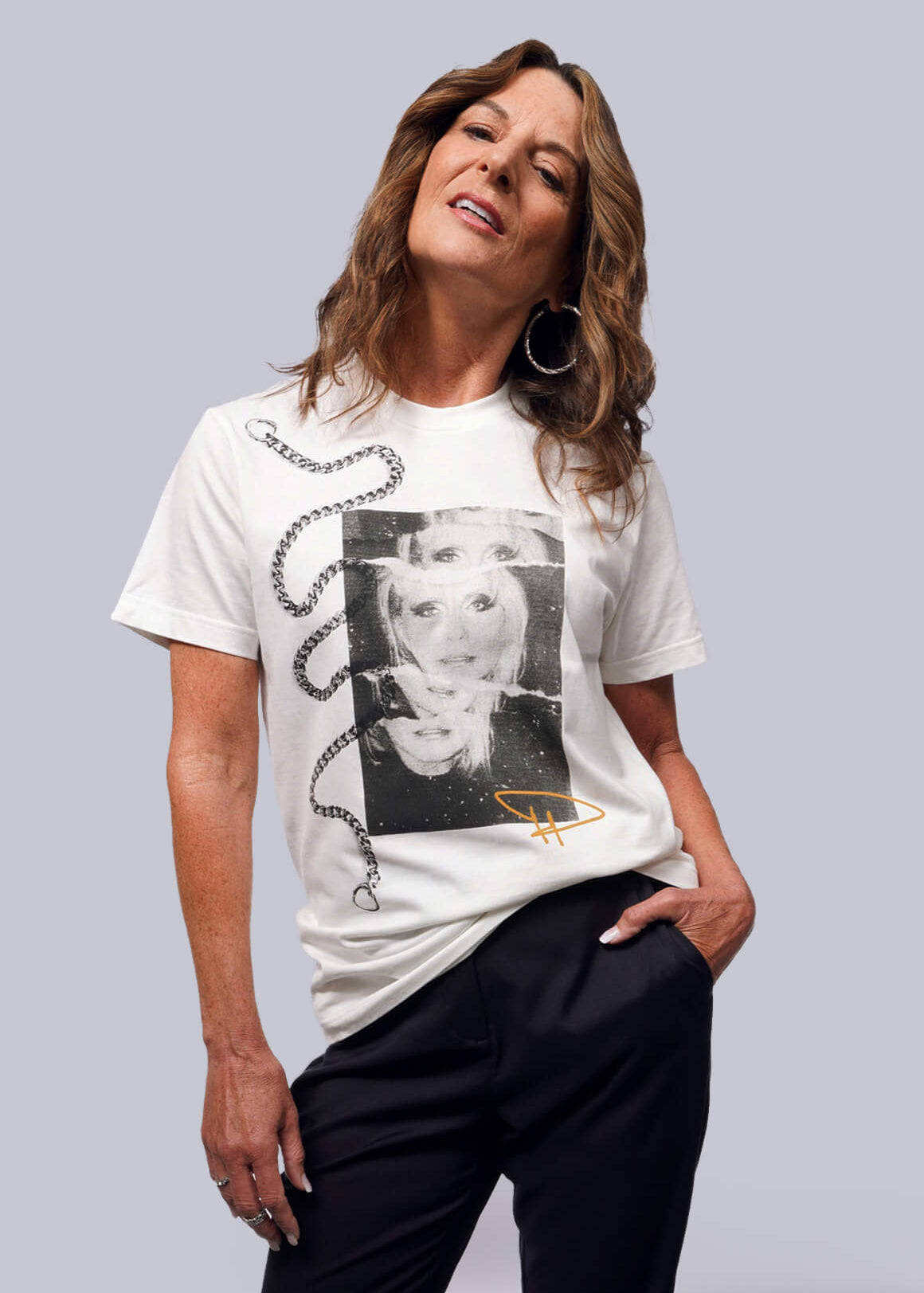S= A person with long hair strikes a confident pose in the Debbie Harry X Wildfang Graphic Tee, showcasing an artistic black-and-white graphic and chain design for a punk vibe. Paired with dark pants, they look upward, hand in pocket, against a plain backdrop.