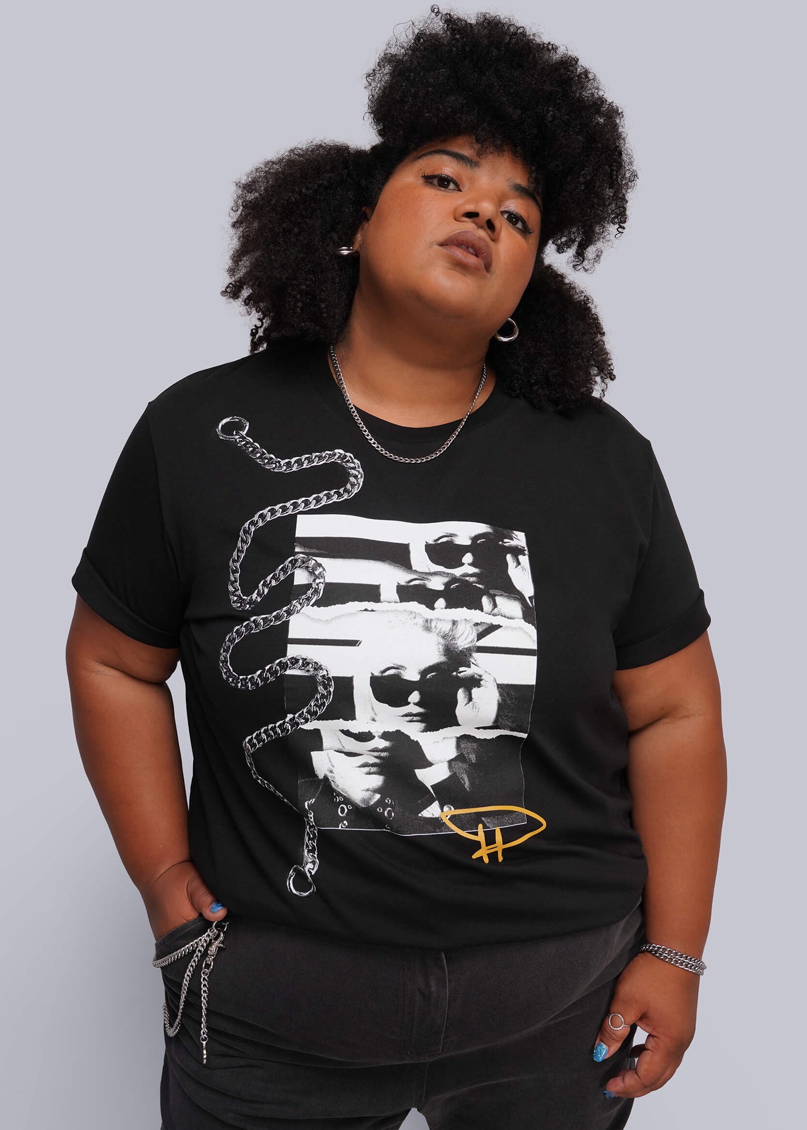 1X=A plus-size model wearing a black graphic t-shirt with a white and yellow print, paired with black jeans, against a solid gray studio background.