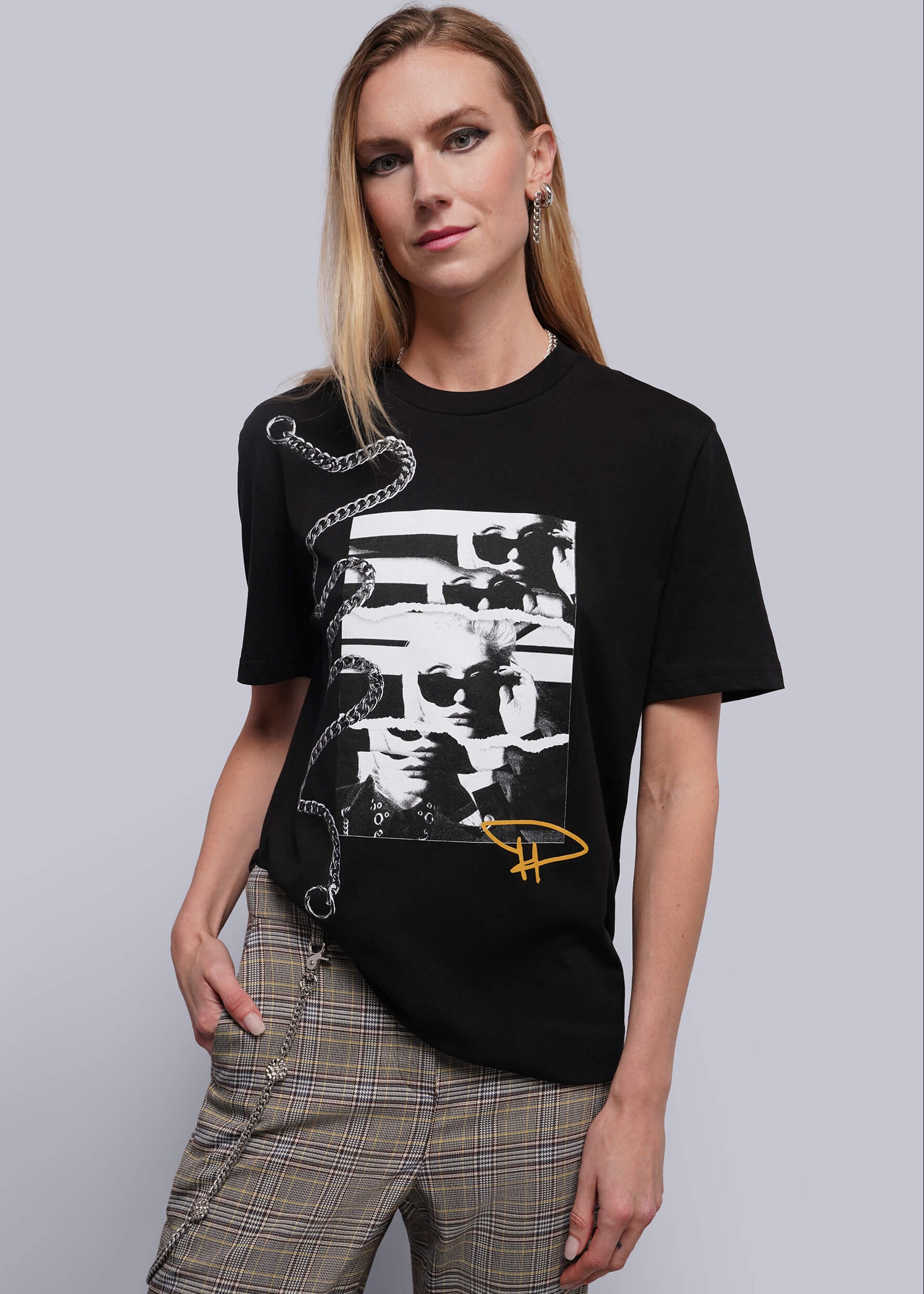 S=A woman modeling a black short-sleeve graphic T-shirt with a large black-and-white photo collage print on the front, a silver chain graphic on the shoulder, and a small yellow signature graphic near the lower right hem, paired with plaid pants against a light gray studio background.