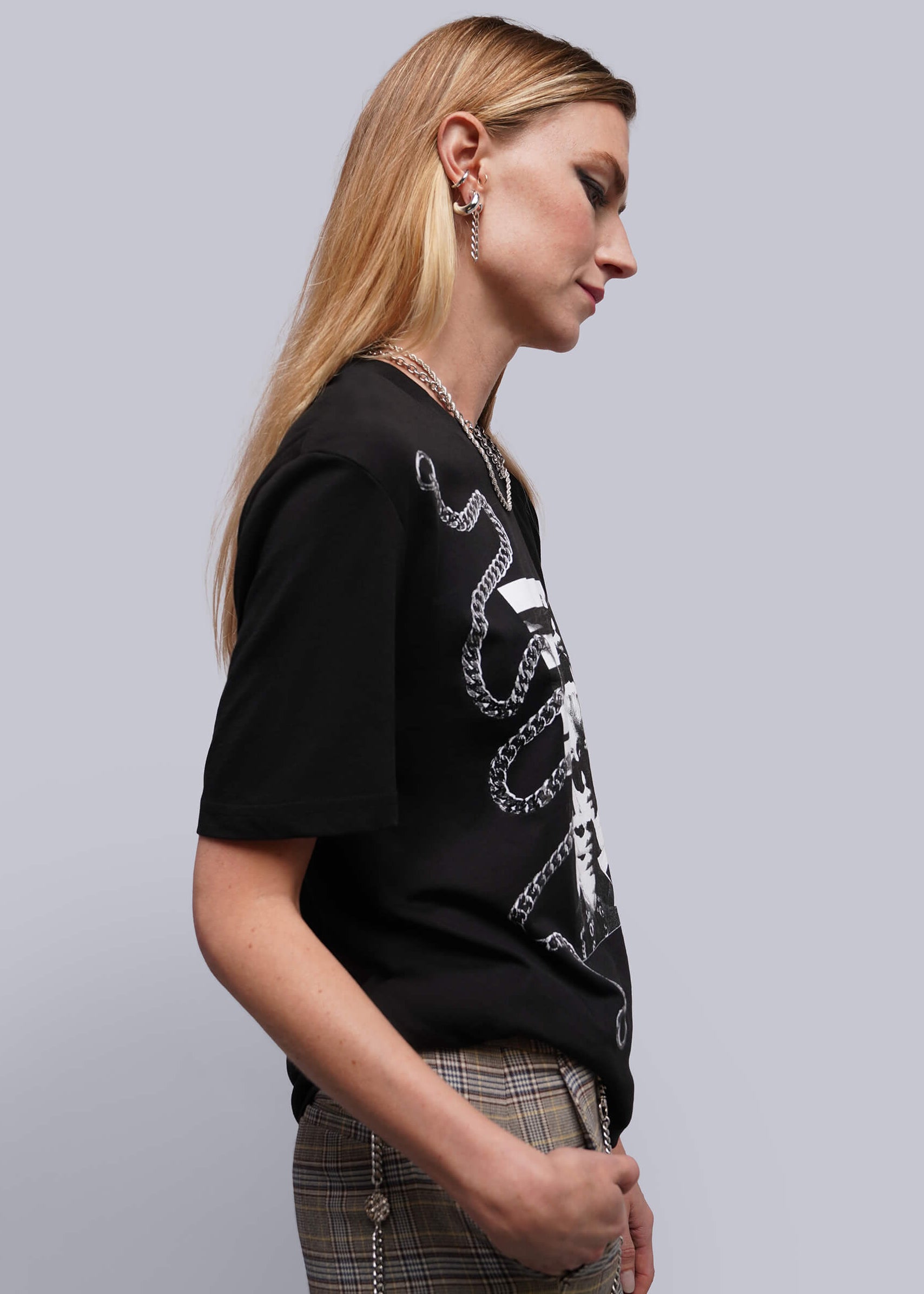 S=A woman in profile wearing a black short-sleeve graphic T-shirt with a white chain print, paired with plaid trousers, against a plain light gray studio background.