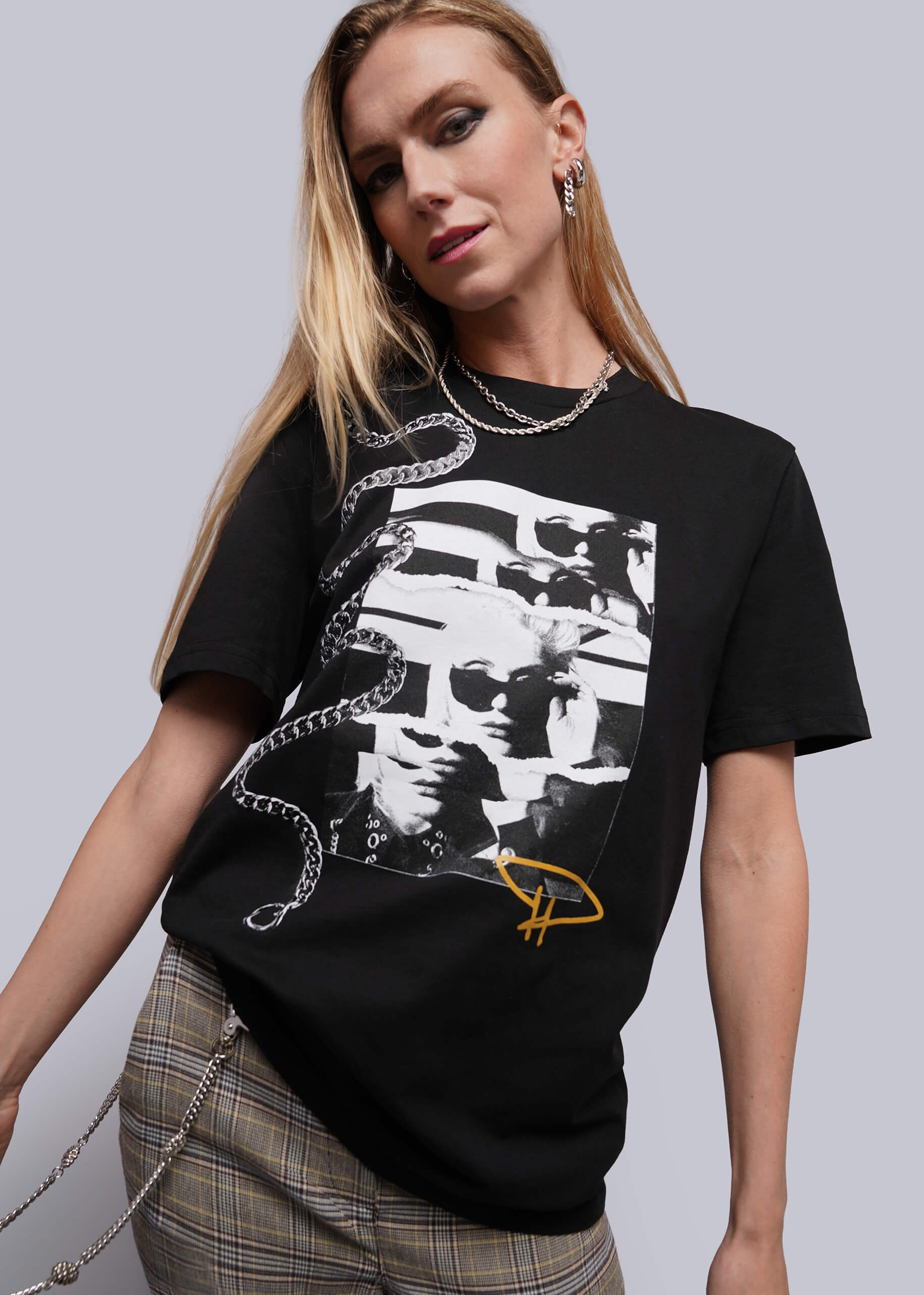 S=A woman modeling a black graphic t-shirt featuring a white and black collage print of a face with chain graphics and a yellow logo, paired with plaid trousers against a solid gray background.