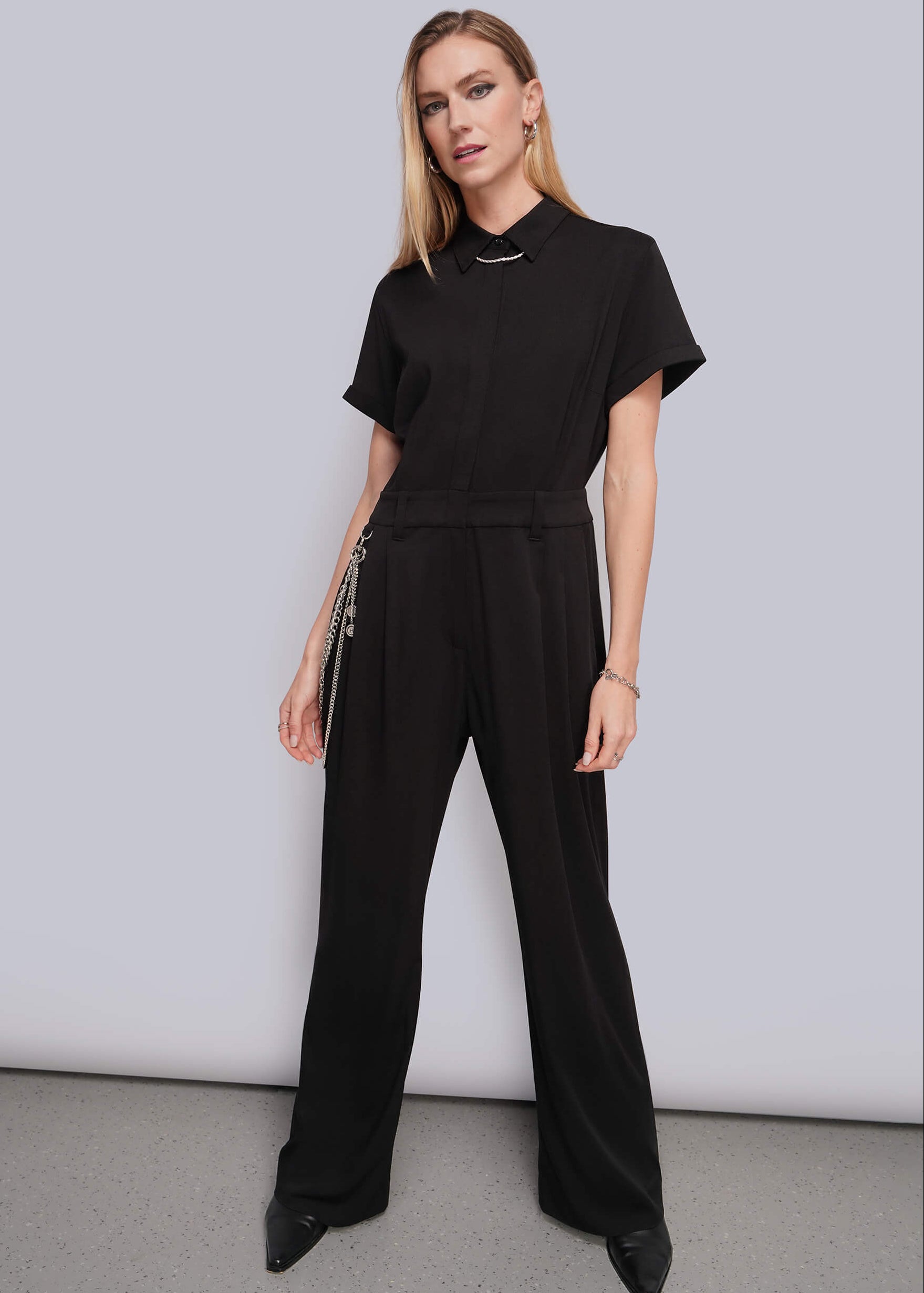 A woman modeling a black short-sleeve jumpsuit with a multi chain wallet chain, standing against a light gray studio background.