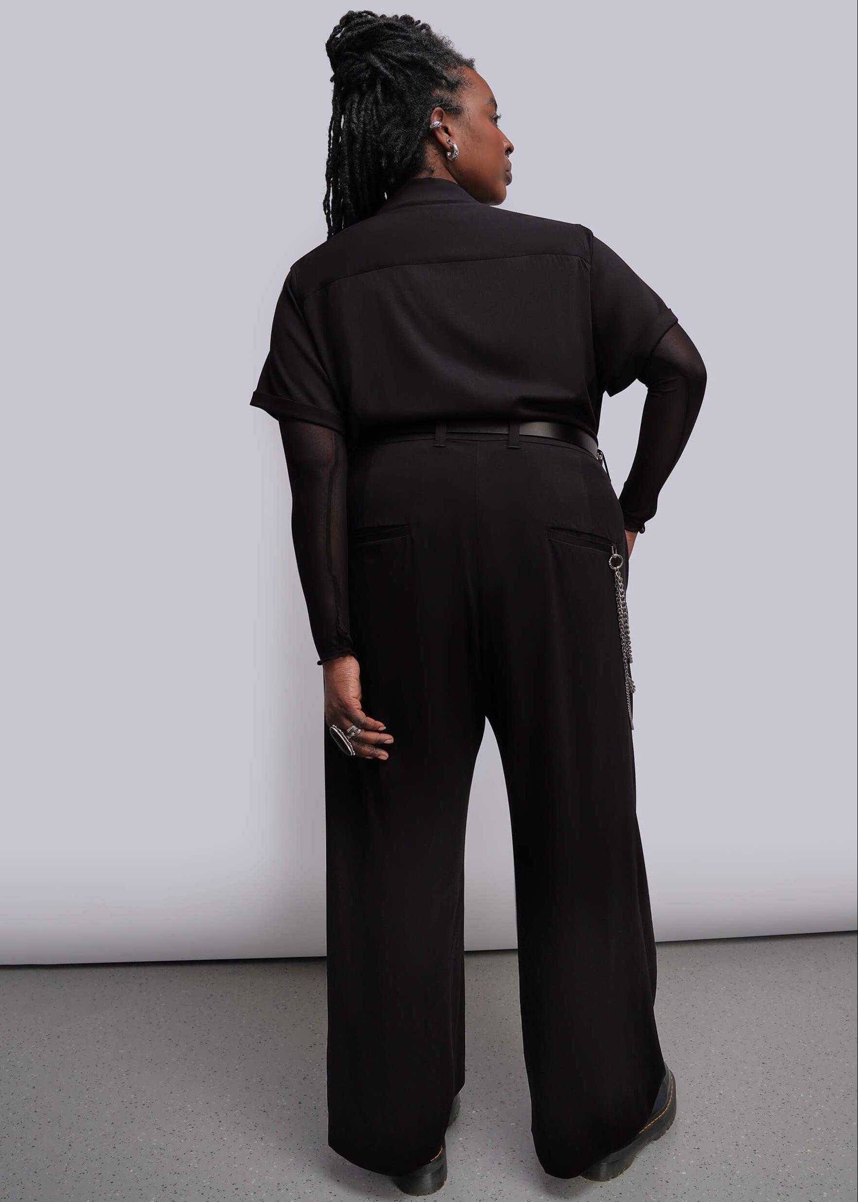 1X=A model shown from the back wearing a black wide-leg jumpsuit with a black belt and a silver wallet chain detail, set against a plain light gray studio background.