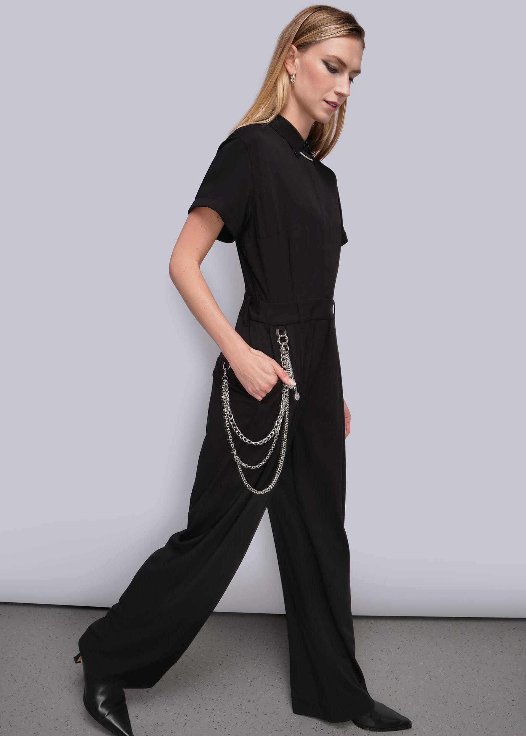 S=A woman modeling a black short-sleeve jumpsuit with wide legs and a collar, styled with layered silver chains draped over her hand, set against a plain light gray studio background.