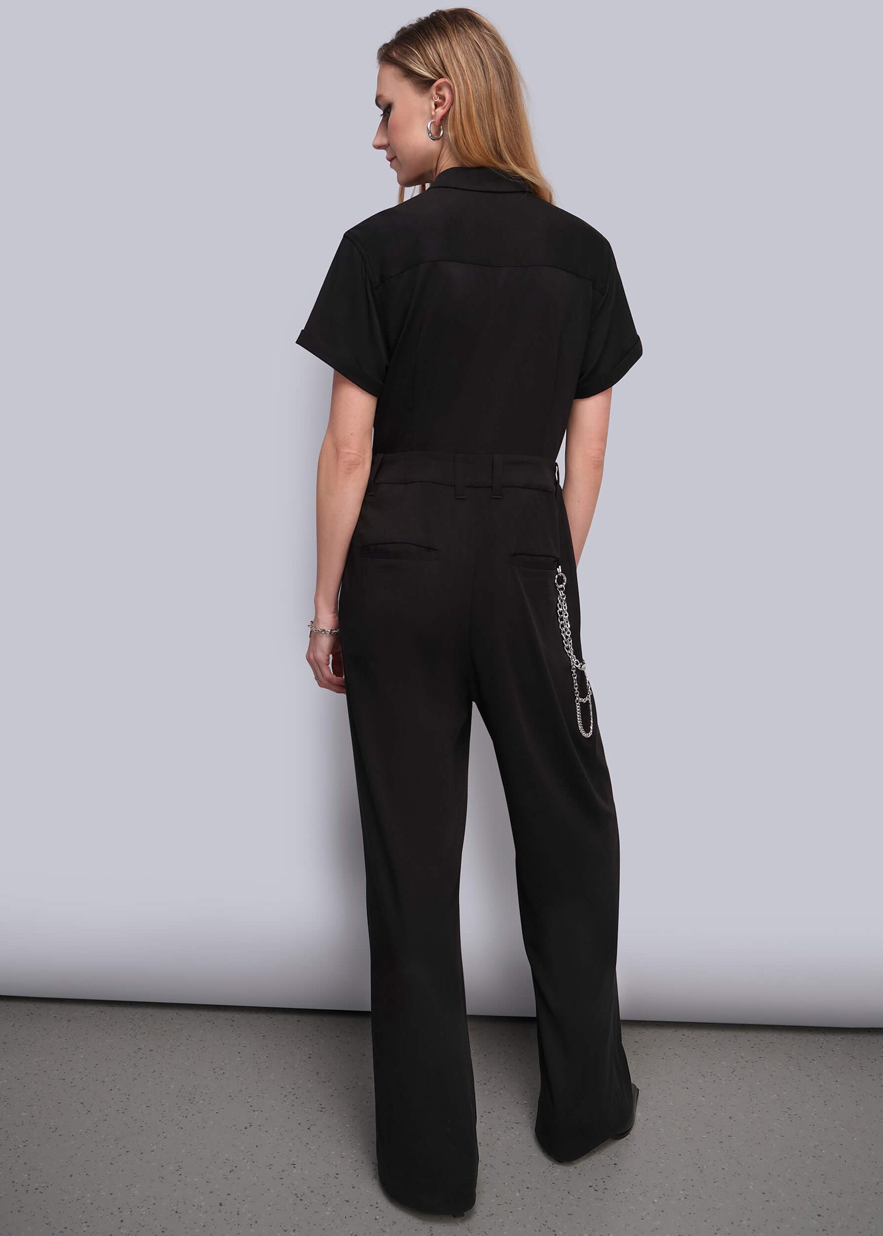 S=A woman shown from the back wearing a black short-sleeve collared jumpsuit with wide legs and a silver chain detail on the side pocket, photographed against a light gray studio background.