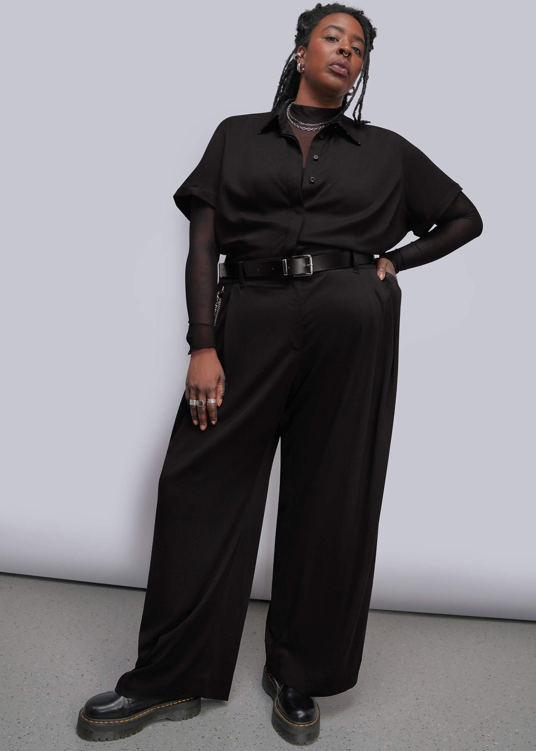 1X=A model wearing a black short-sleeve jumpsuit with a sheer mesh top underneath, a black belt with a silver buckle, and black platform shoes, standing against a light gray studio background.