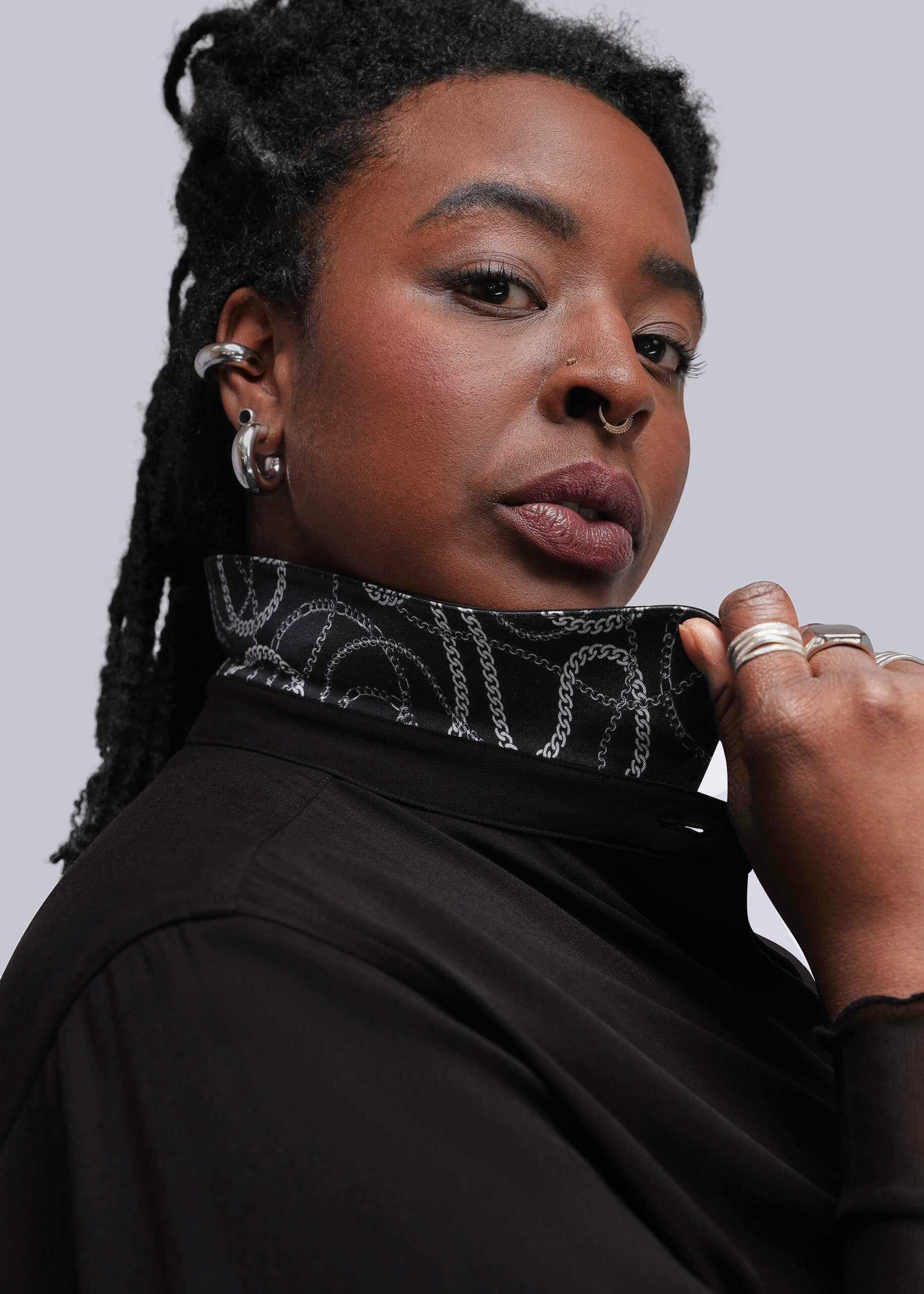 1X=A close-up portrait of a woman holding up collar of jumpsuit showing pattern underneath, set against a solid light gray background.