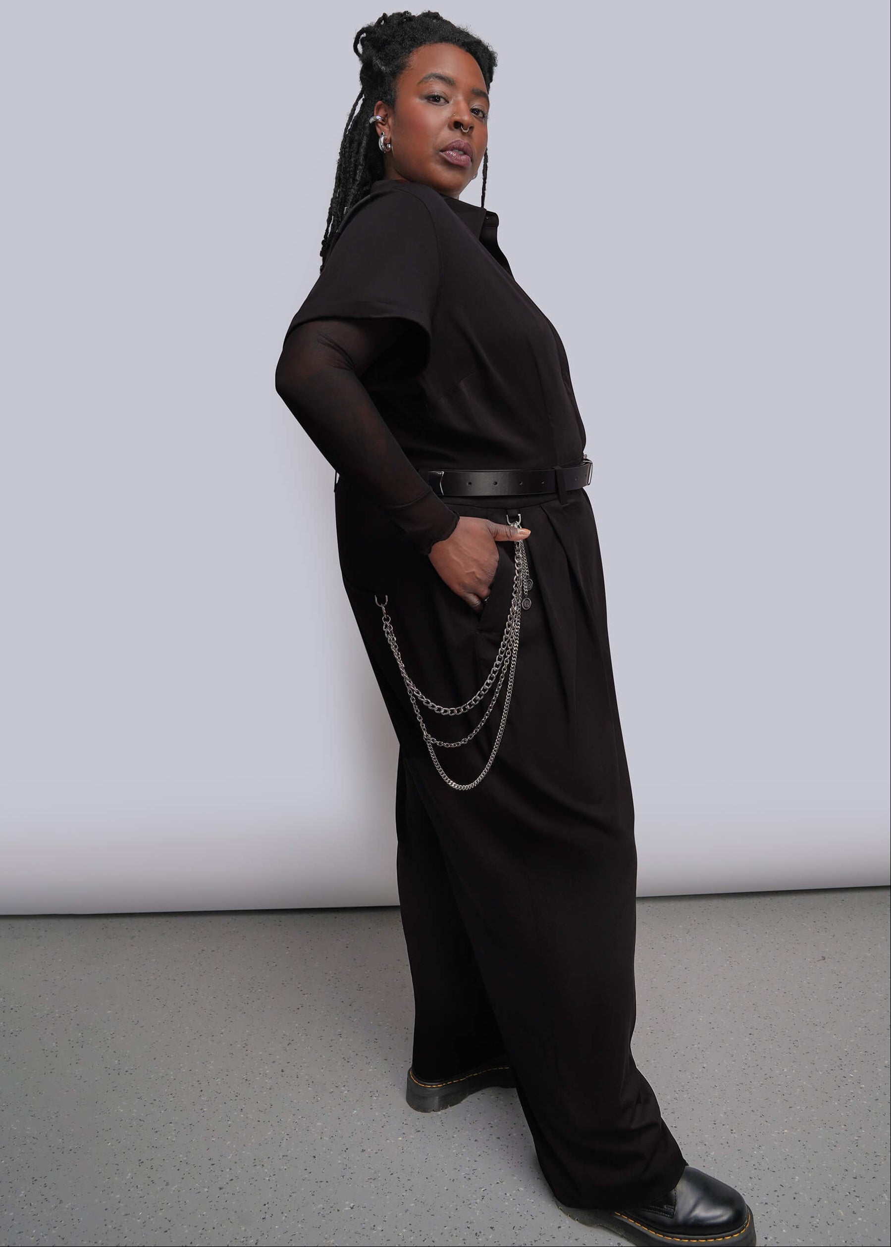 1X=A model wearing a black short sleeve wide leg jumpsuit and a sheer mesh long sleeve top underneath, styled with a black belt and layered silver chains, standing against a light gray studio backdrop.