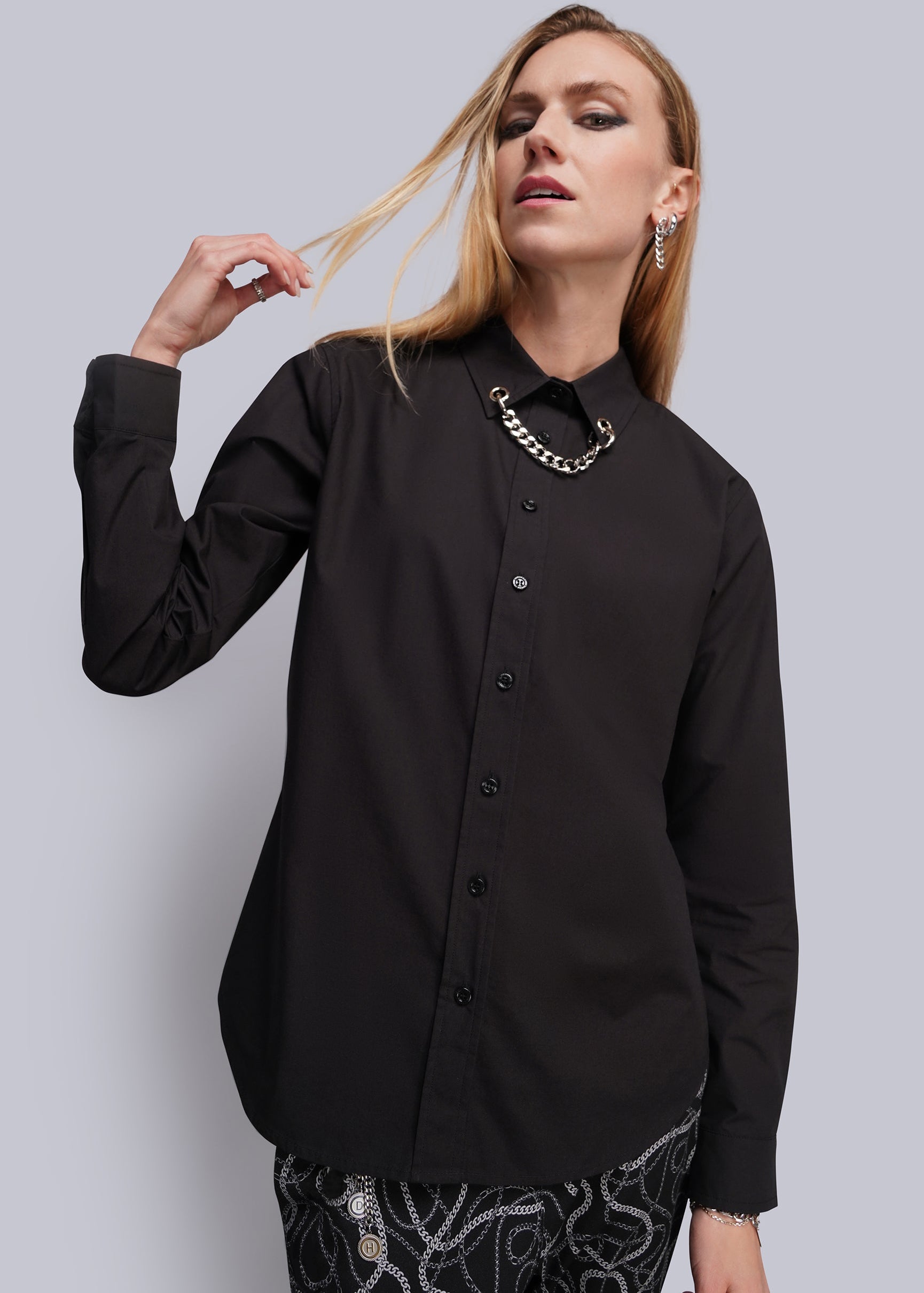 A woman modeling a black long-sleeve button-up shirt with a grommet and chain embellished collar, paired with black and white patterned pants, against a solid gray studio background.
