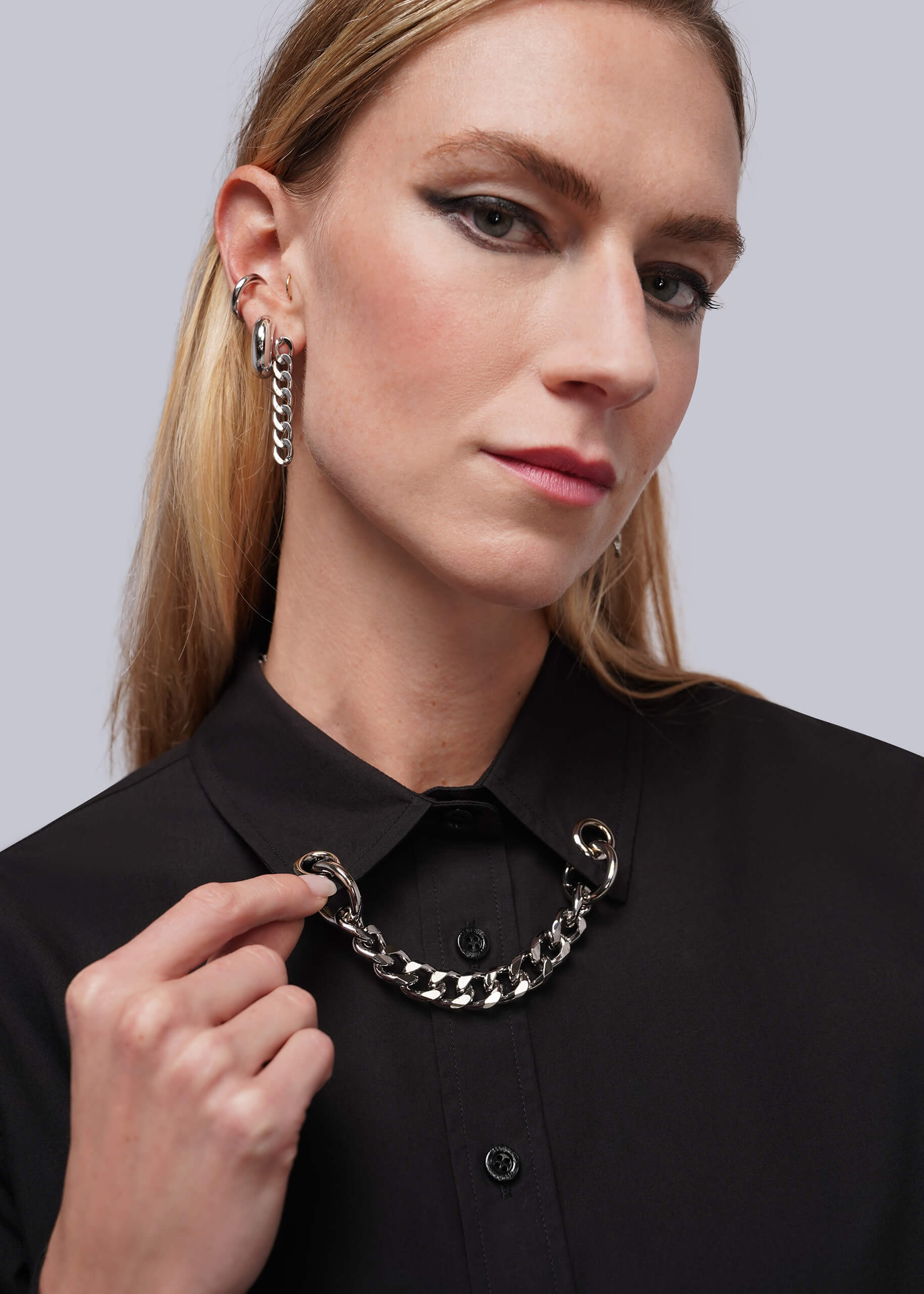 A close-up of a woman wearing a black button up shirt with a grommet and chain embellished collar and silver earrings.