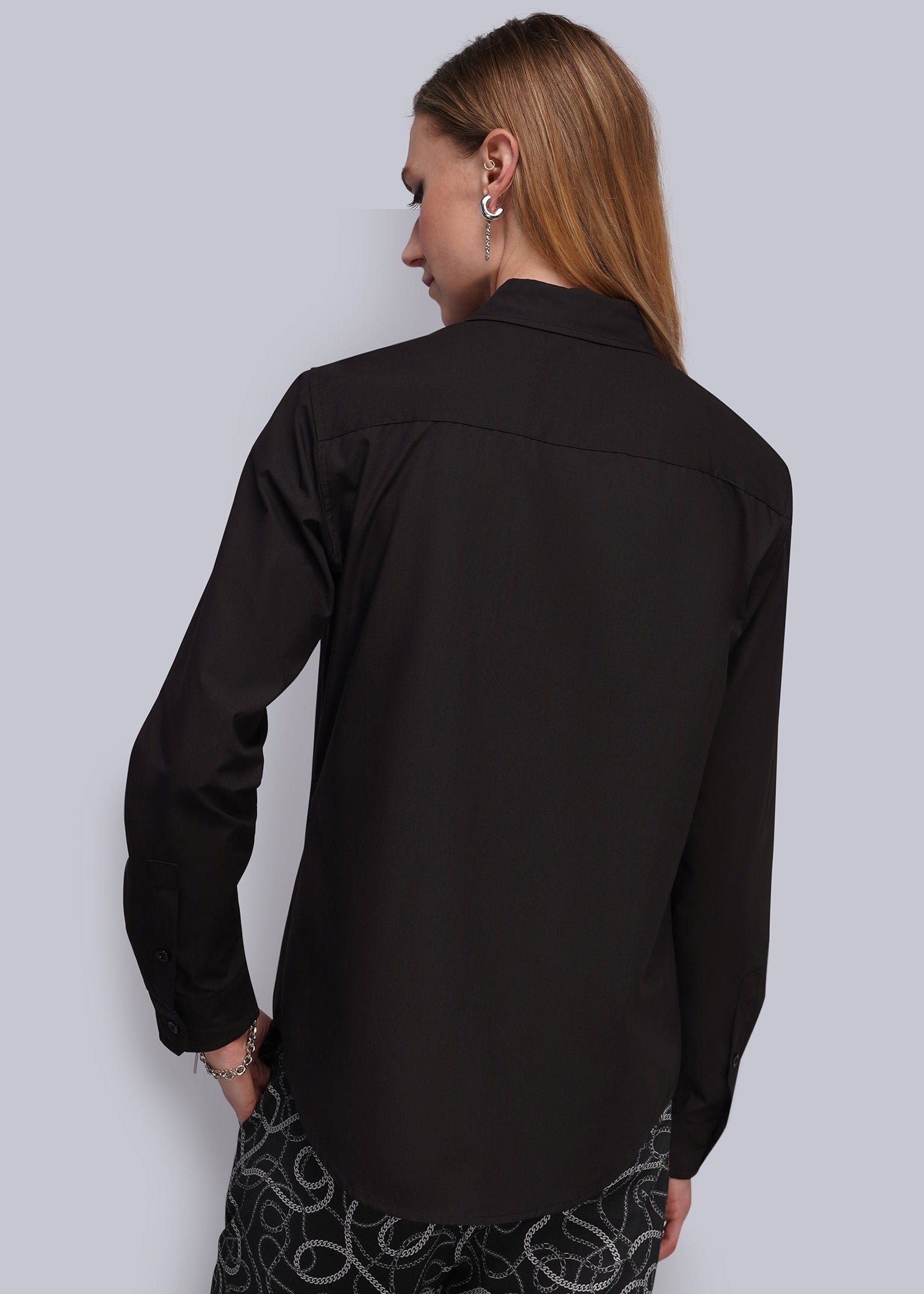 S=A woman shown from the back wearing a black long-sleeve button-up shirt and black pants with a white abstract pattern, set against a solid light gray background.