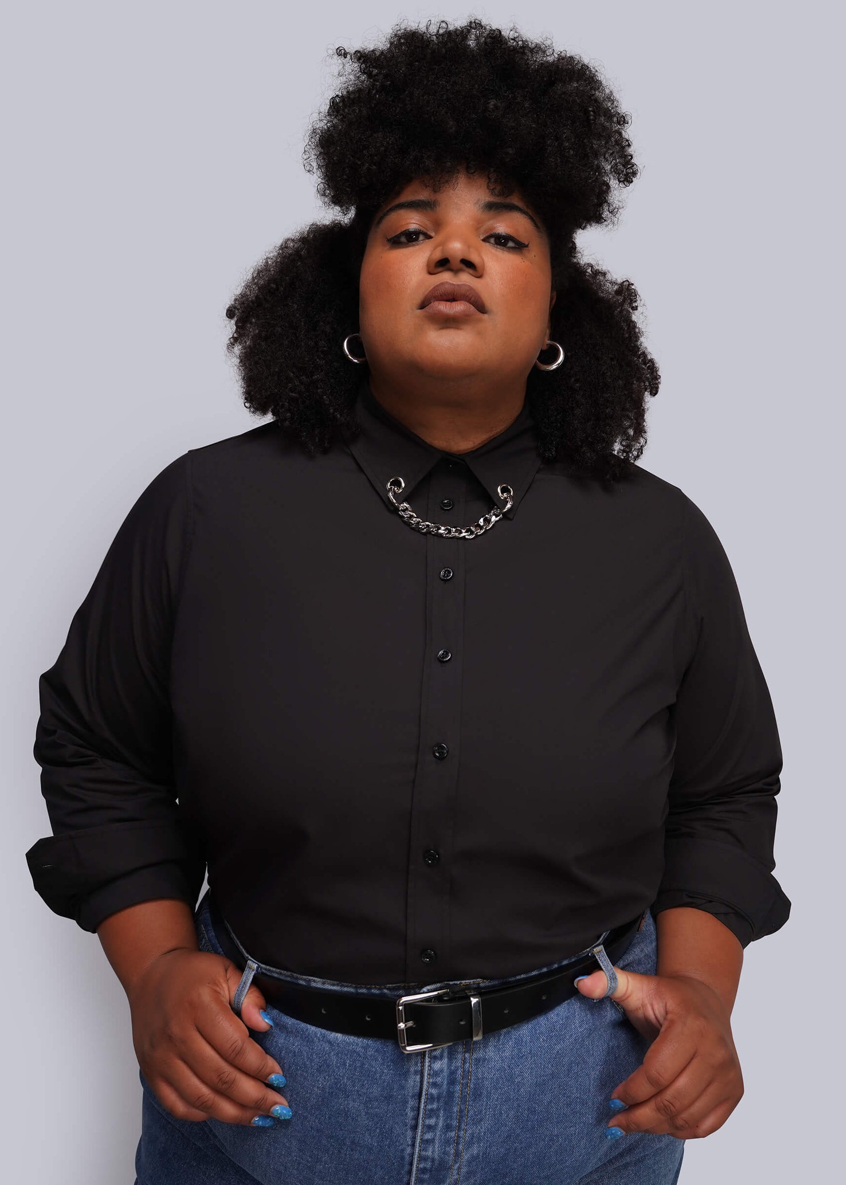 1X=A person wearing a black long-sleeve button-up shirt with a grommet and chain embellished collar, blue jeans, and a black belt, standing against a solid light gray background.