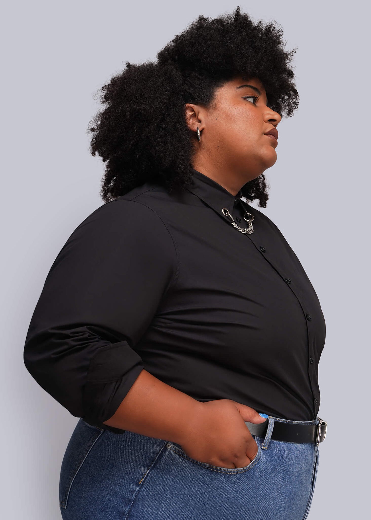 1X=A woman with curly black hair is shown in profile wearing a black long-sleeve button-up shirt and blue jeans against a solid light gray background.