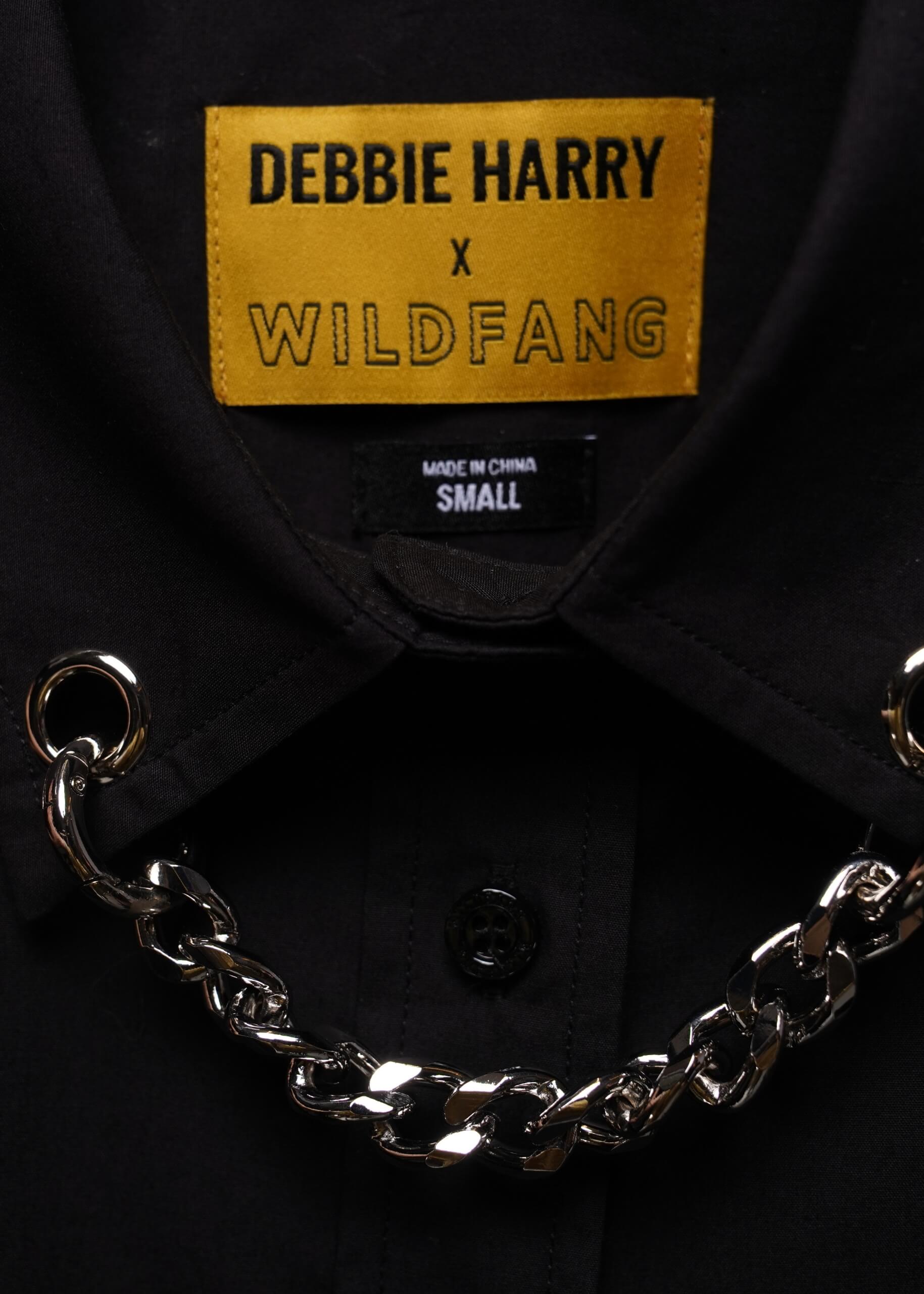 Close-up of a black collared shirt with a gold label reading 'DEBBIE HARRY x WILDFANG' and a chunky silver chain draped across the collar area.