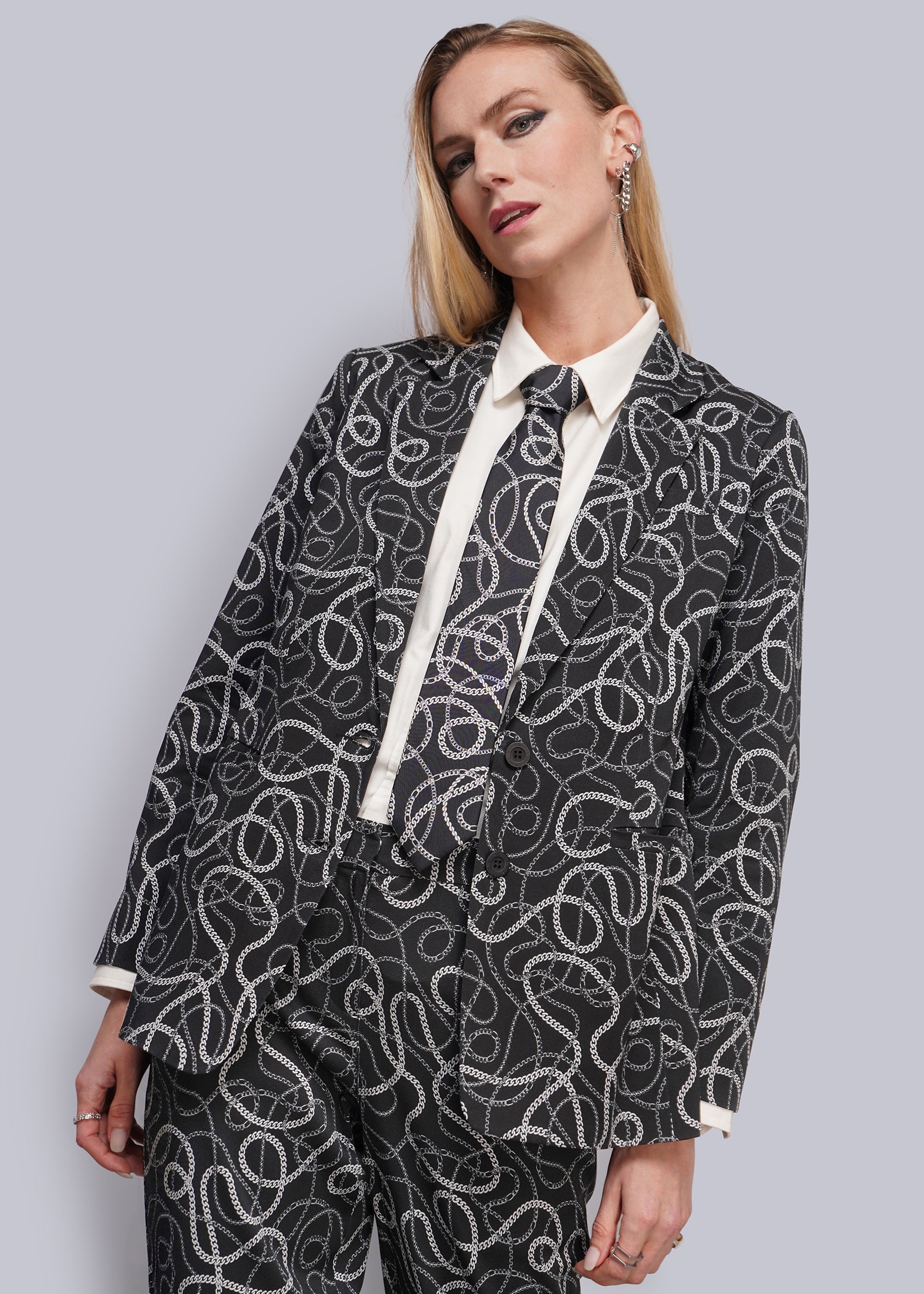 A woman modeling a black and white abstract patterned blazer and matching trousers, styled with a white collared shirt and tie, against a solid gray studio background.