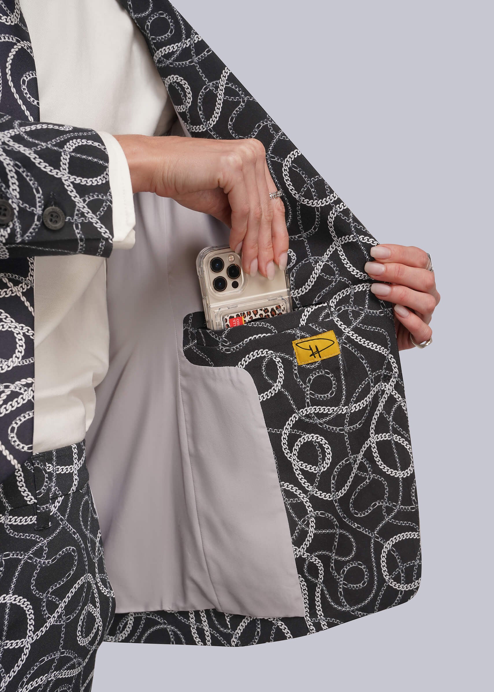 A close-up of a person wearing a black blazer with a white chain-link pattern, holding the lapel open to show a light gray interior pocket containing a smartphone and a small yellow tag.