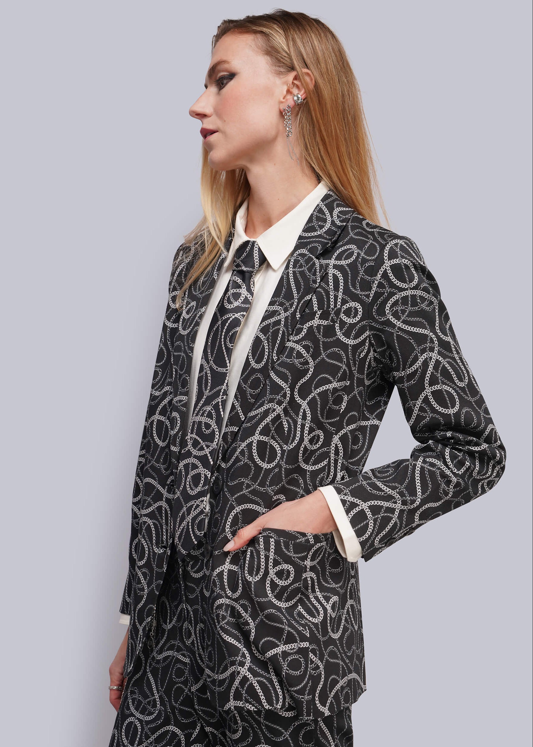S=A woman shown in profile wearing a black blazer with a white abstract chain pattern, layered over a white collared shirt. She is photographed against a solid light gray studio background.