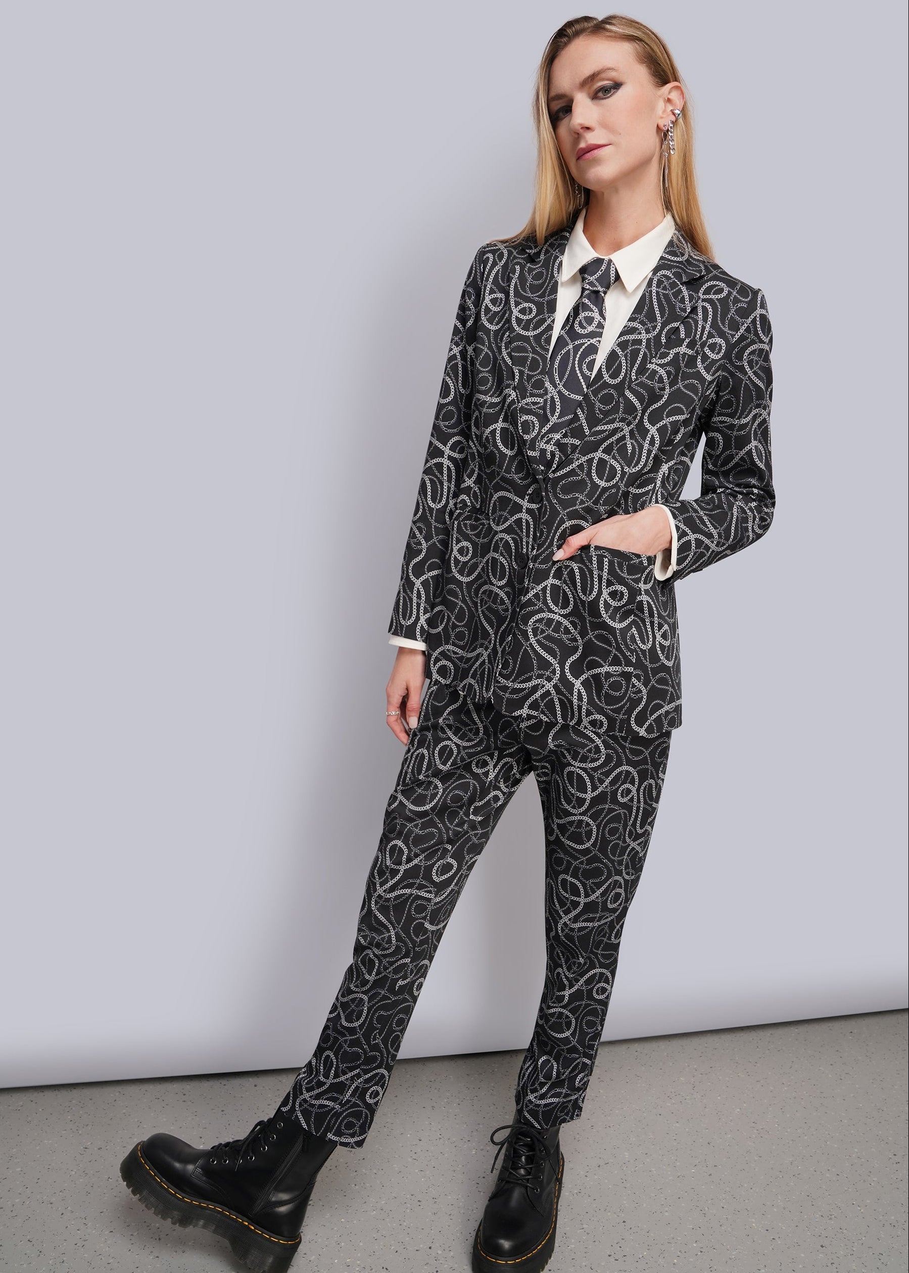 S=A woman modeling a black and white chain patterned blazer and matching trousers suit, styled with a white collared shirt and black leather boots against a light gray studio background.