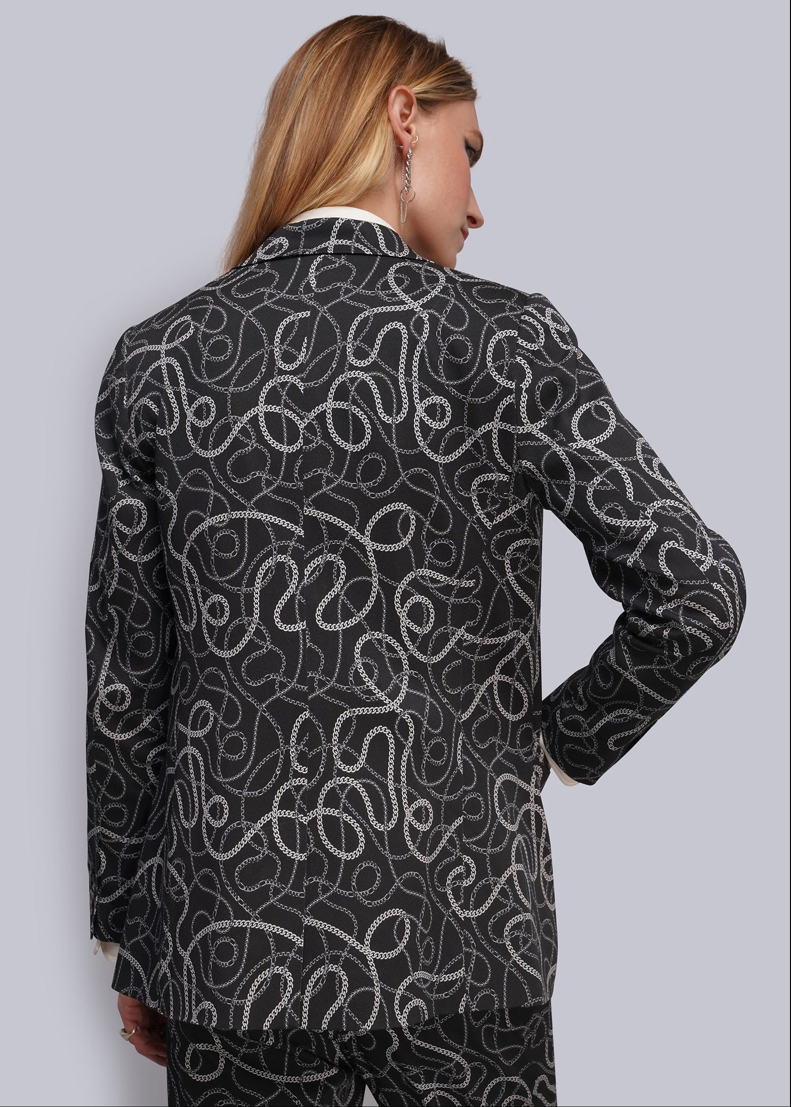 S=A woman shown from the back wearing a black blazer and matching trousers with an all-over white and grey wavy chain pattern, against a light gray studio background.