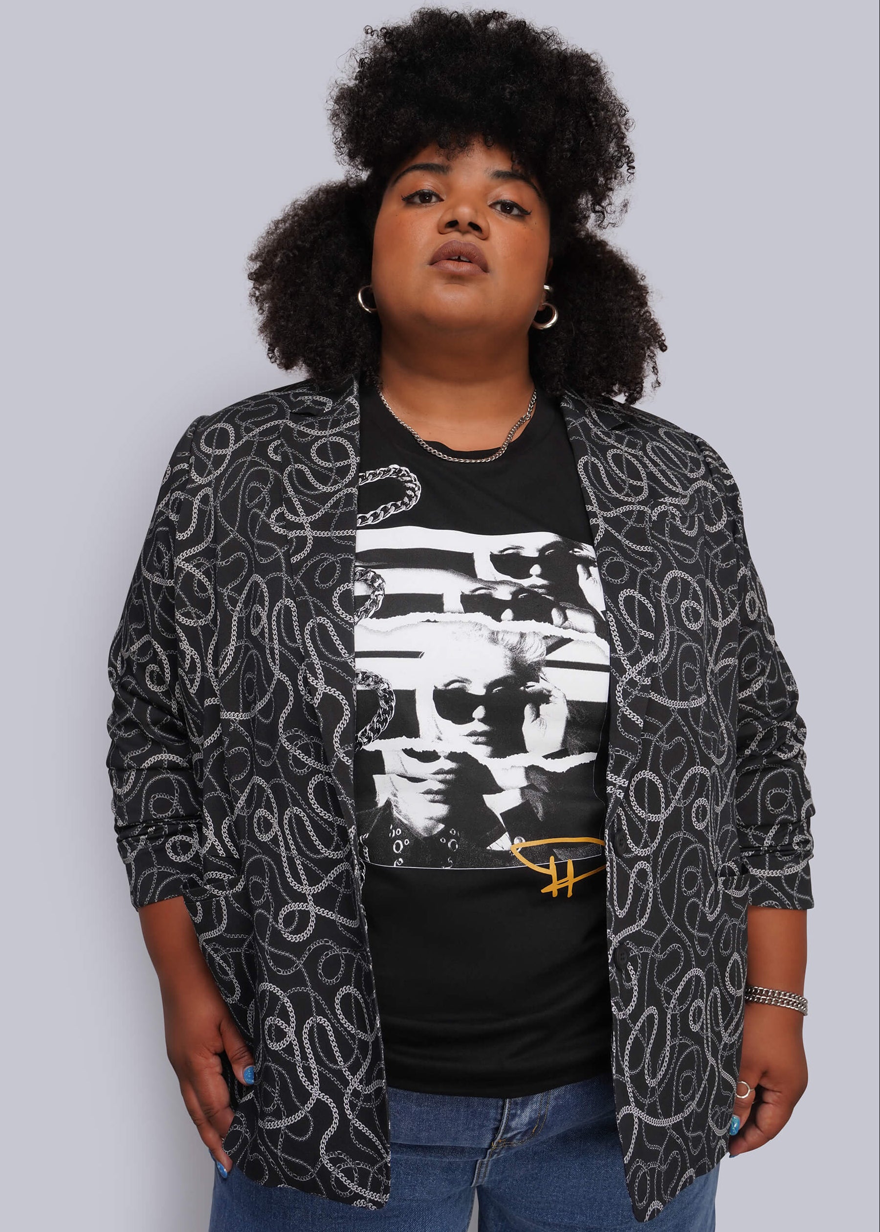 1X=A person wearing a black graphic T-shirt under an open black-and-white chain patterned blazer, paired with blue jeans, against a plain light gray studio background.