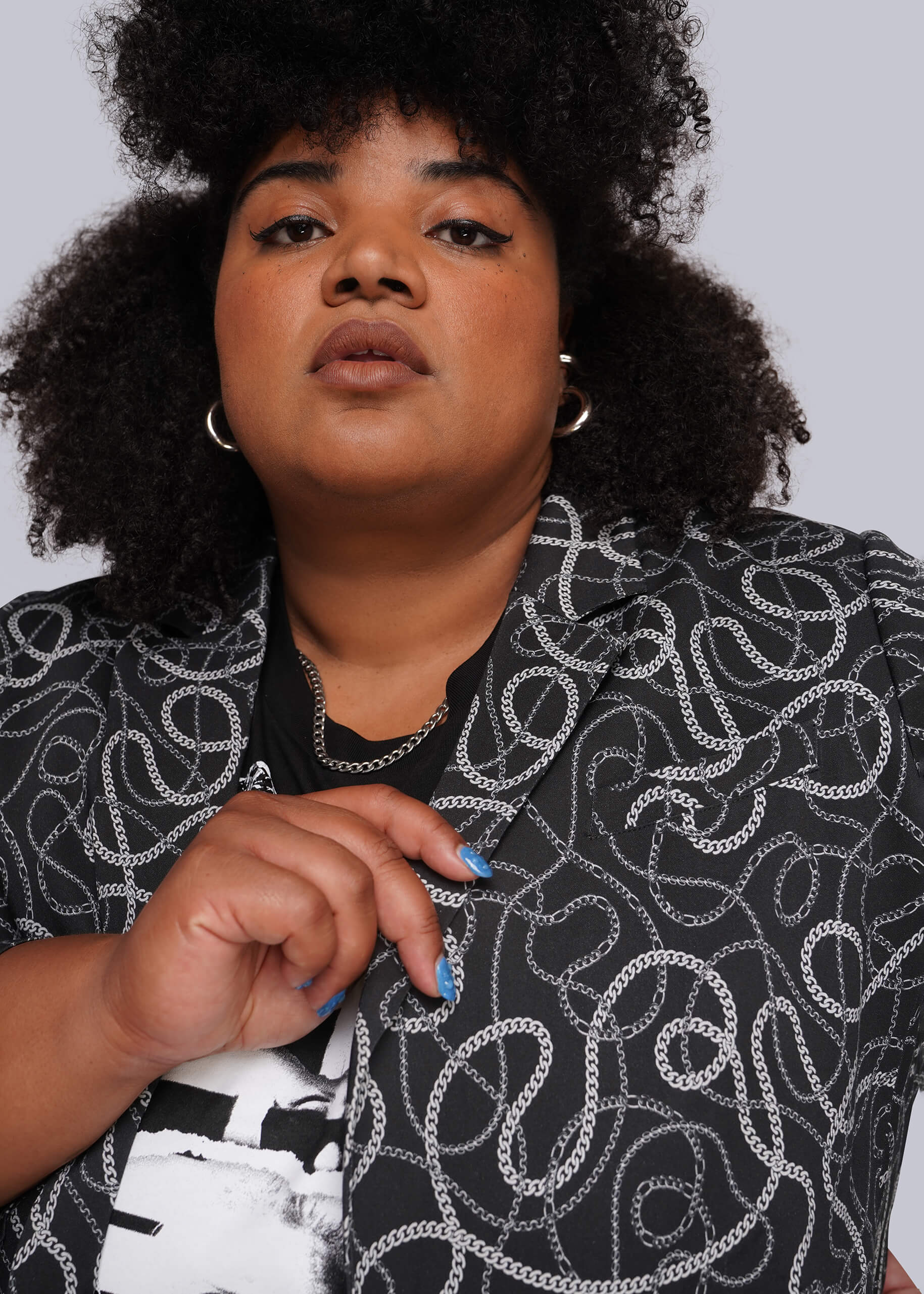 1X=A close-up portrait of a model wearing a black blazer with a white and grey chain-link pattern and a black-and-white graphic t-shirt underneath.