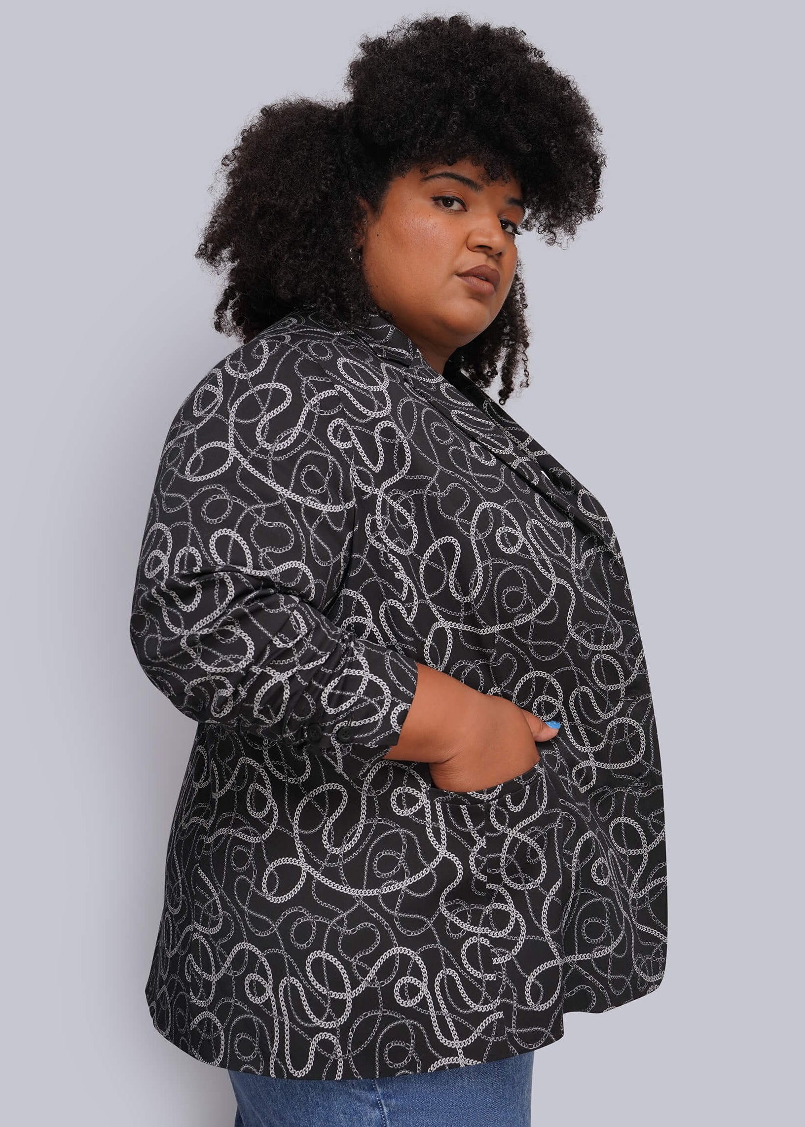 1X=A woman with dark curly hair models a black blazer featuring a white and grey chain-link pattern, paired with blue jeans, against a solid light gray background.