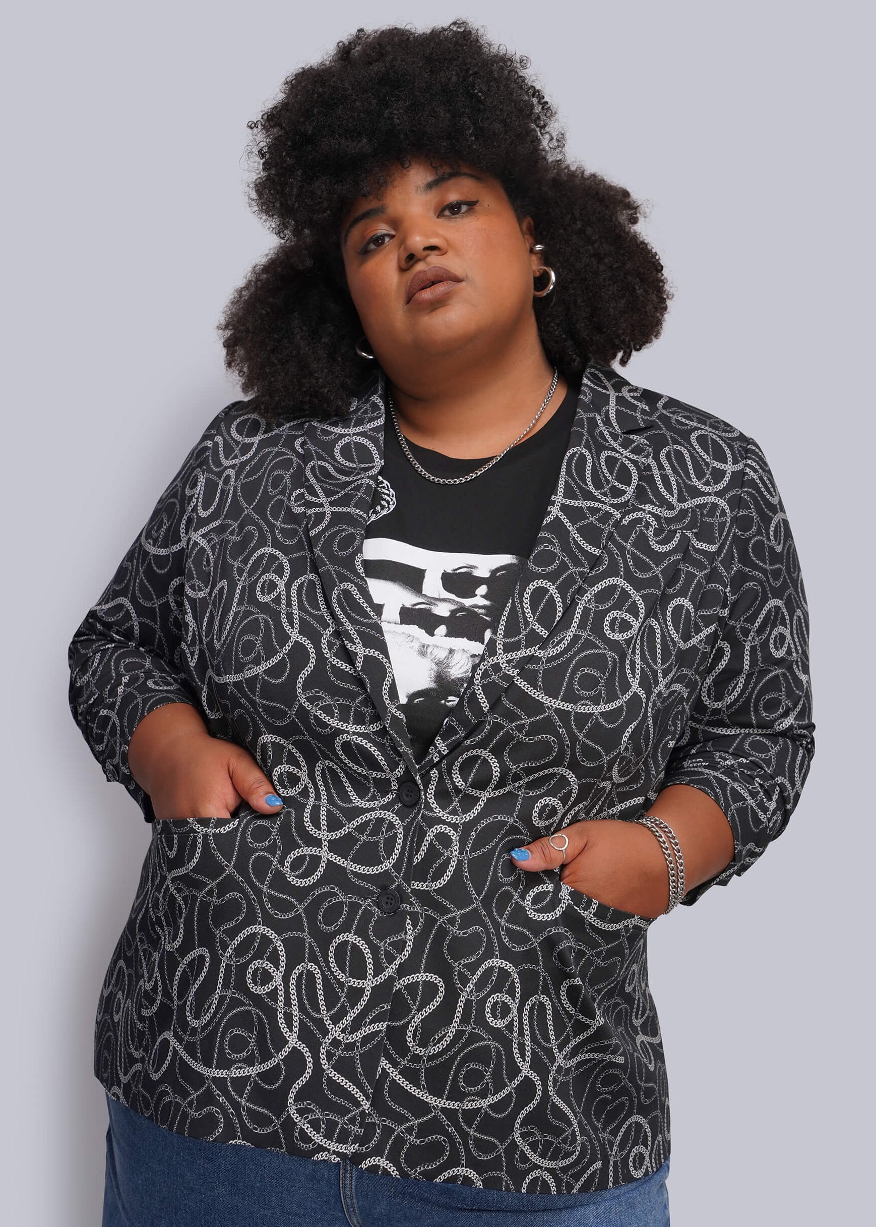 1X=A plus-size model wearing a black and white chain-link patterned blazer over a black graphic t-shirt and blue jeans, standing against a solid light gray background.