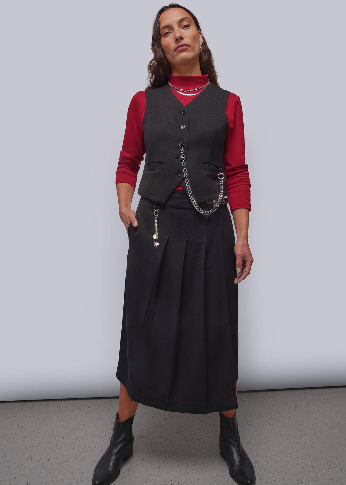 S=A model stands against a light gray studio background wearing a black pleated midi skirt and matching vest with a silver chain detail, layered over a red long-sleeve top and black ankle boots.
