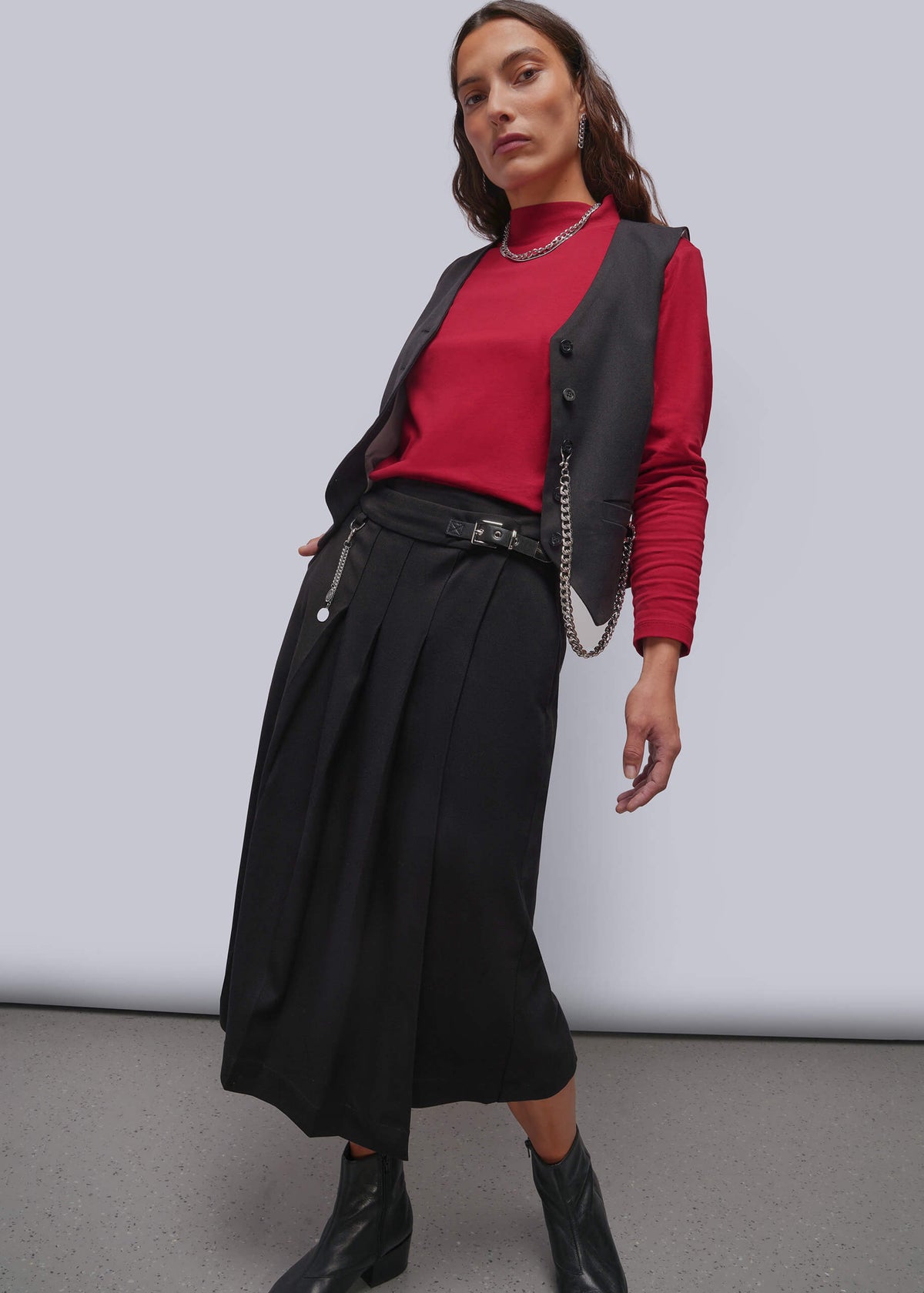 S=A model wearing a black pleated midi skirt with a silver zipper and chain detail, styled with a red long-sleeve top, a black vest, and black ankle boots against a light gray studio background.