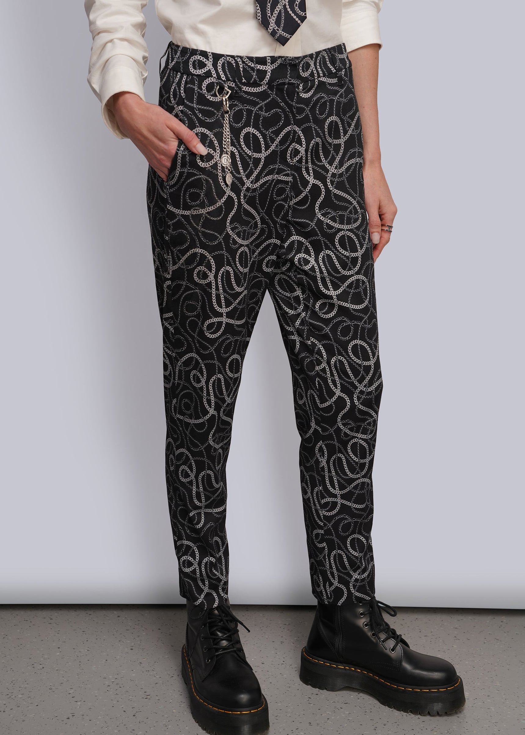 A person wearing black pants with a white and grey chain-link wavy pattern, styled with a cream shirt, a patterned tie, and silver chain accessories, paired with black leather boots.