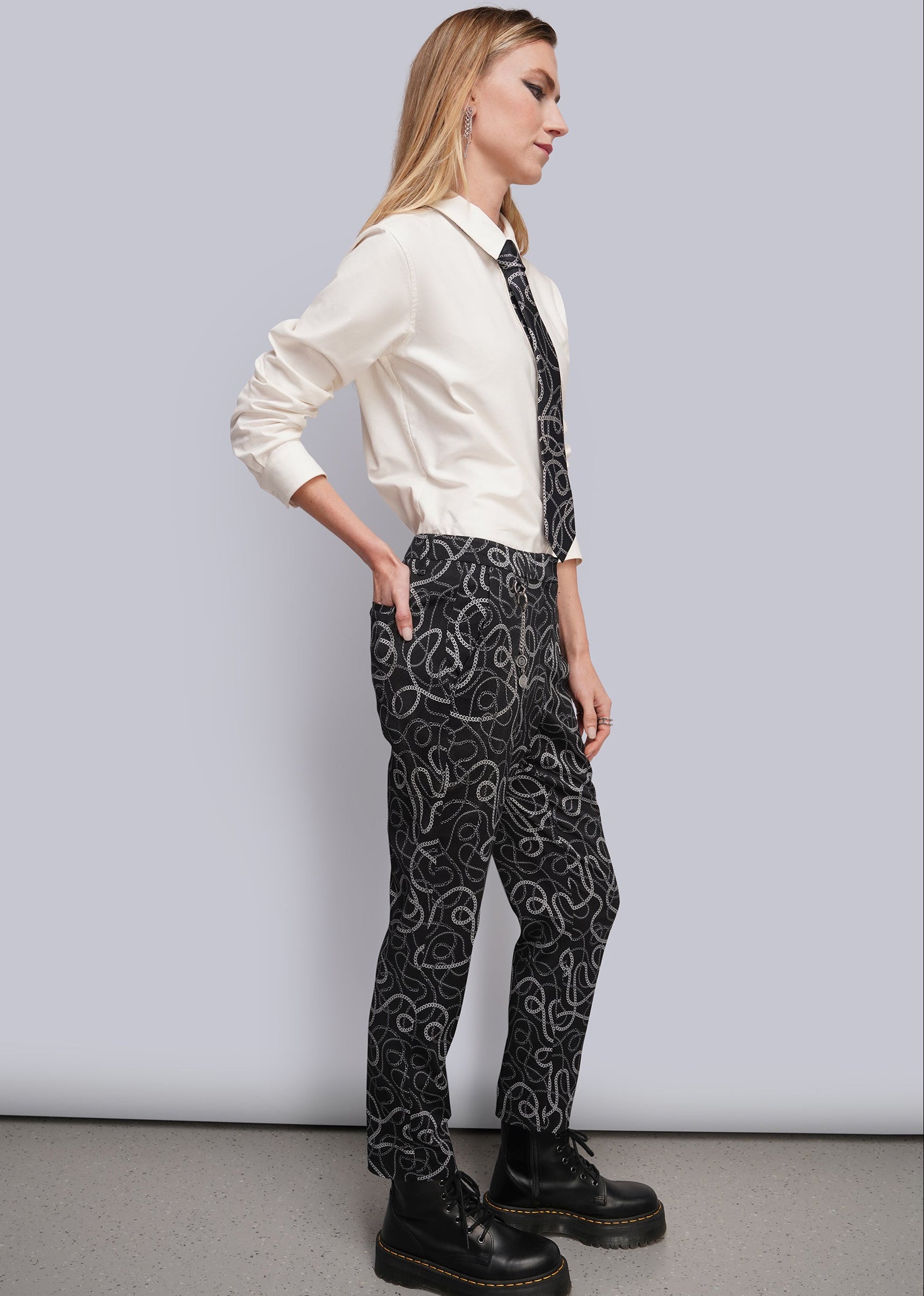 S=A woman stands in profile wearing a cream long-sleeve collared shirt, a black necktie with a chain-link pattern, and black trousers with a matching chain-link print and draped chain details. She is wearing black platform lace-up boots against a light gray studio background.