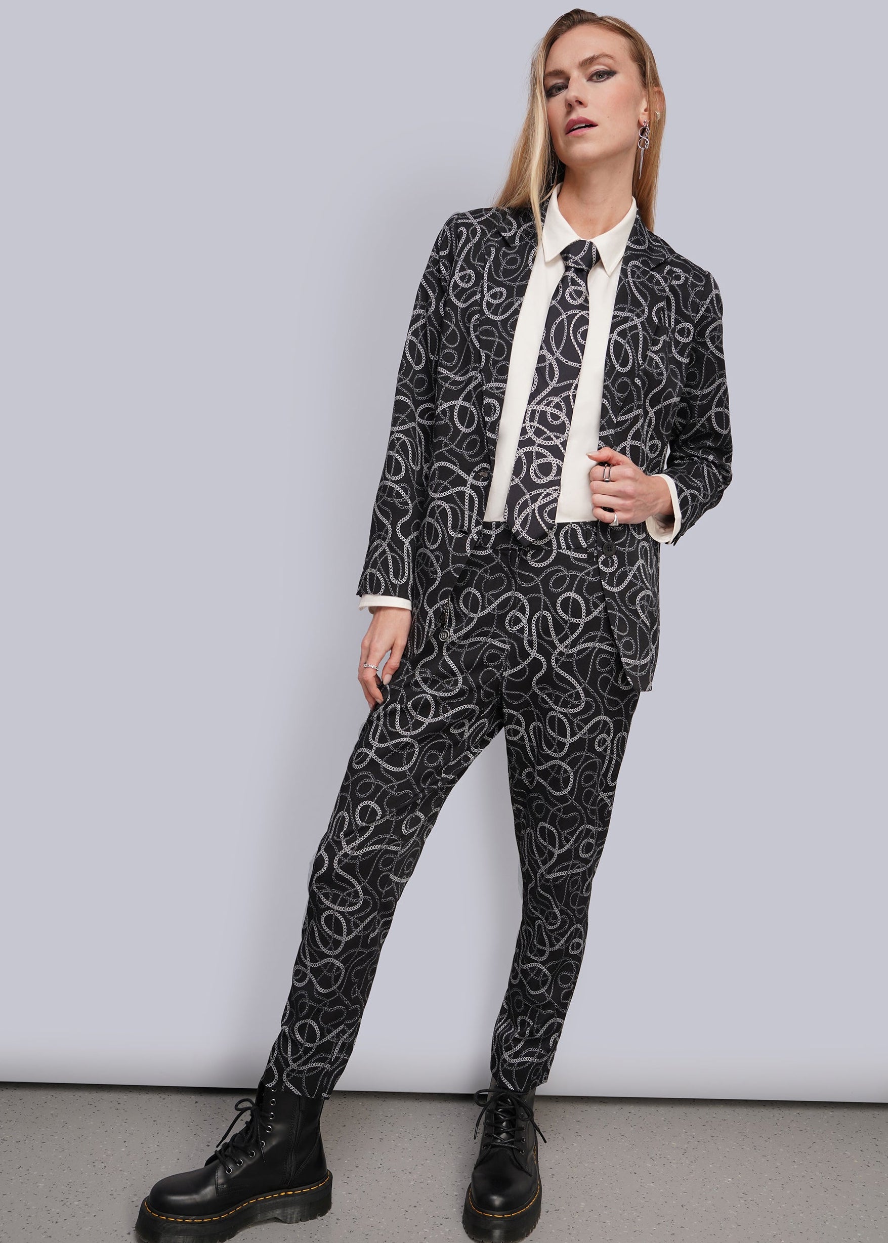 A woman modeling a black and white chain-link patterned suit with a white shirt and black boots, standing against a light gray studio background.