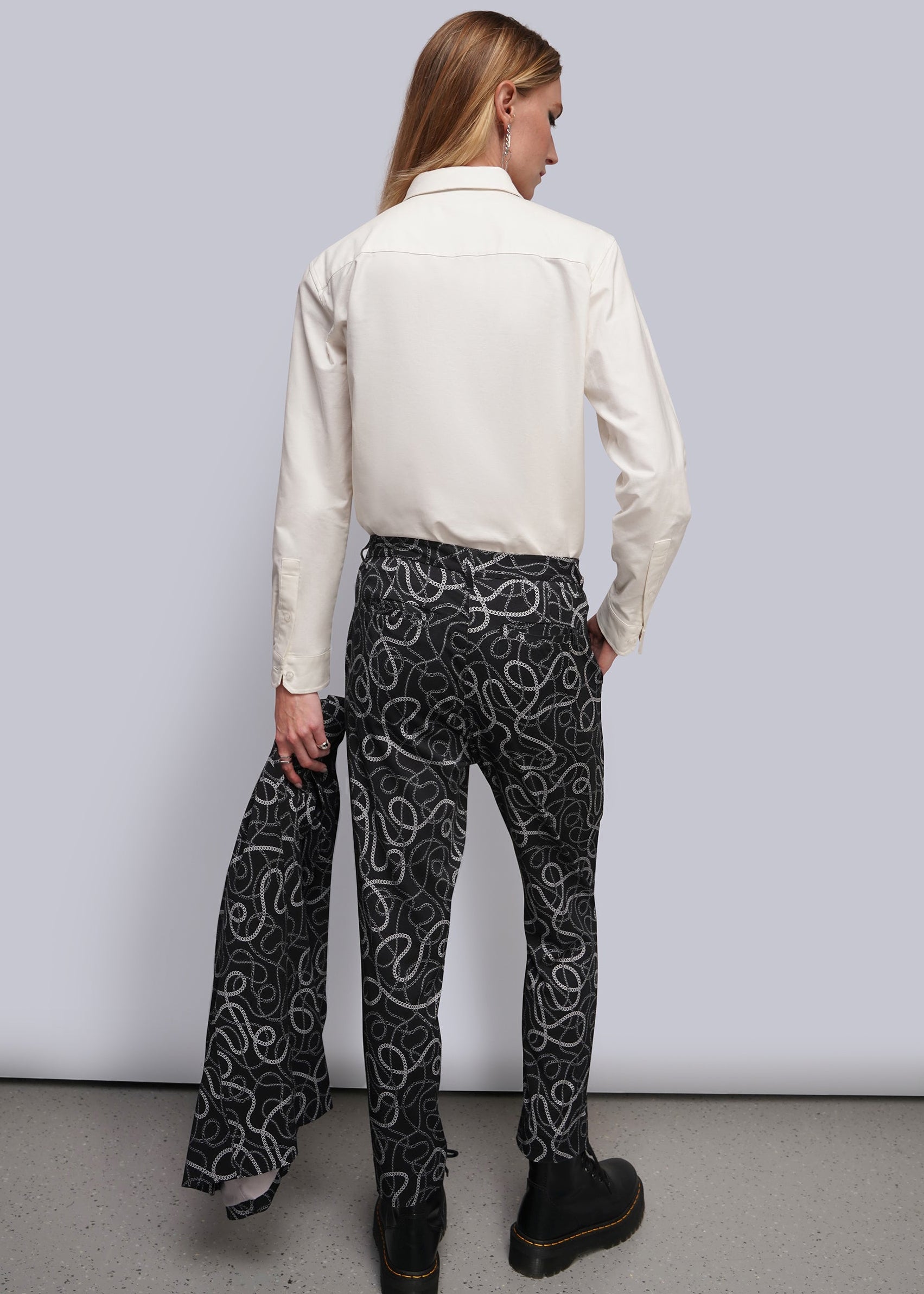 S=A model shown from the back wearing a cream long-sleeve shirt and black trousers with a white chain-link pattern, standing against a light gray studio background.