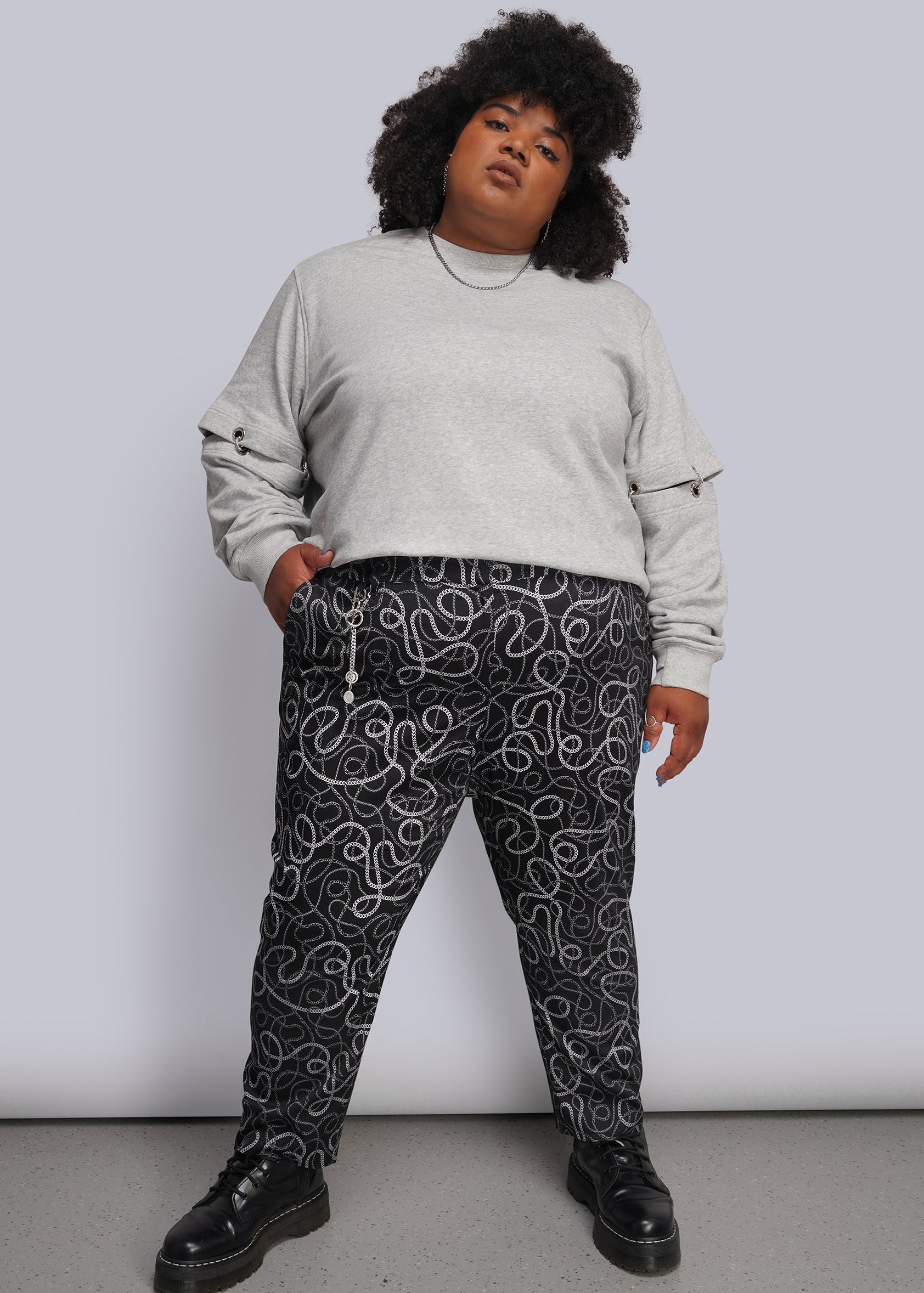 1X=A person modeling a light gray crewneck sweatshirt and black pants with a white and grey chain-link pattern, paired with black boots, against a plain light gray studio background.