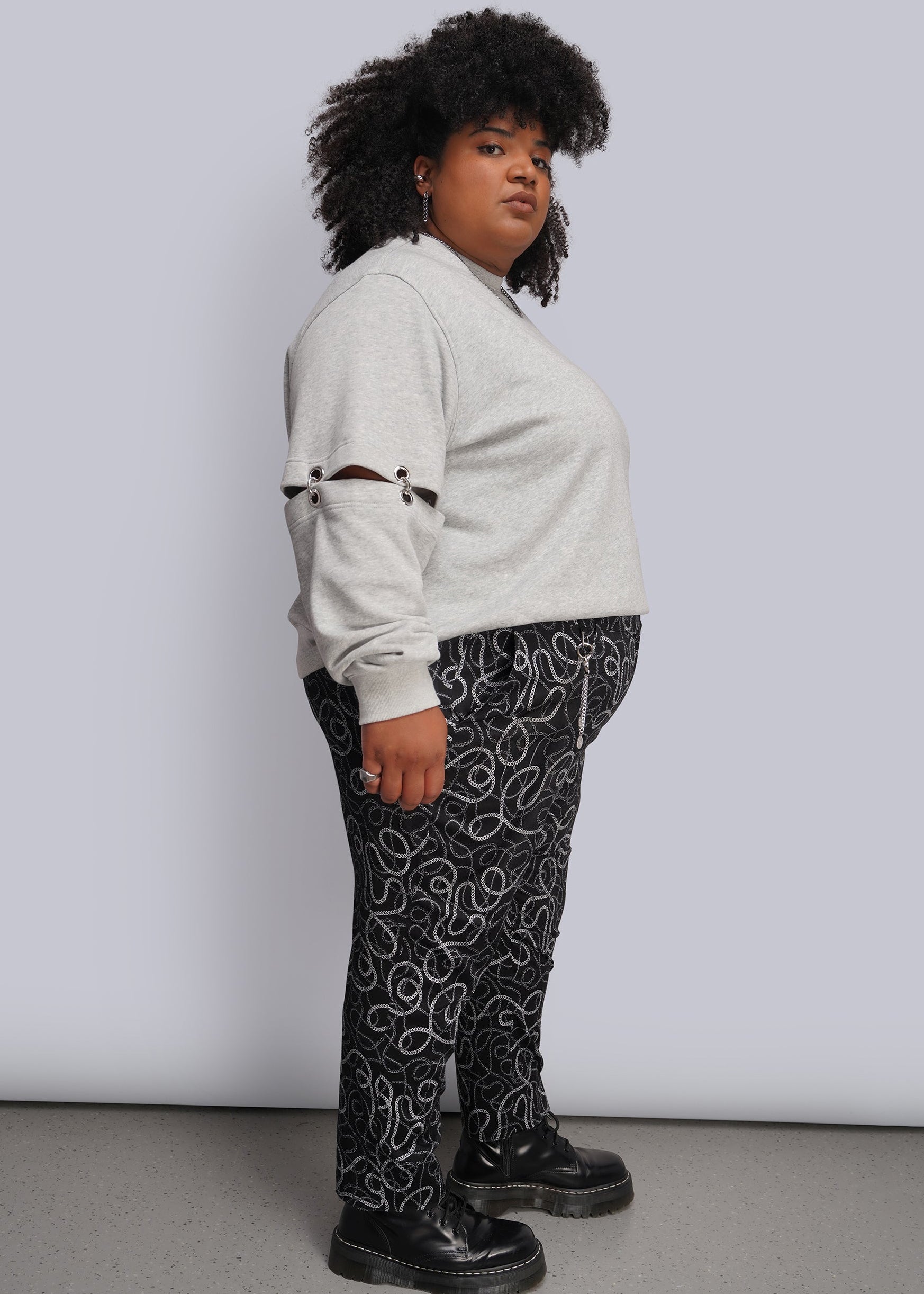 1X=A person stands in profile wearing a light gray sweatshirt, black-and-white chain-link print pants, and black platform shoes against a plain light gray studio background.