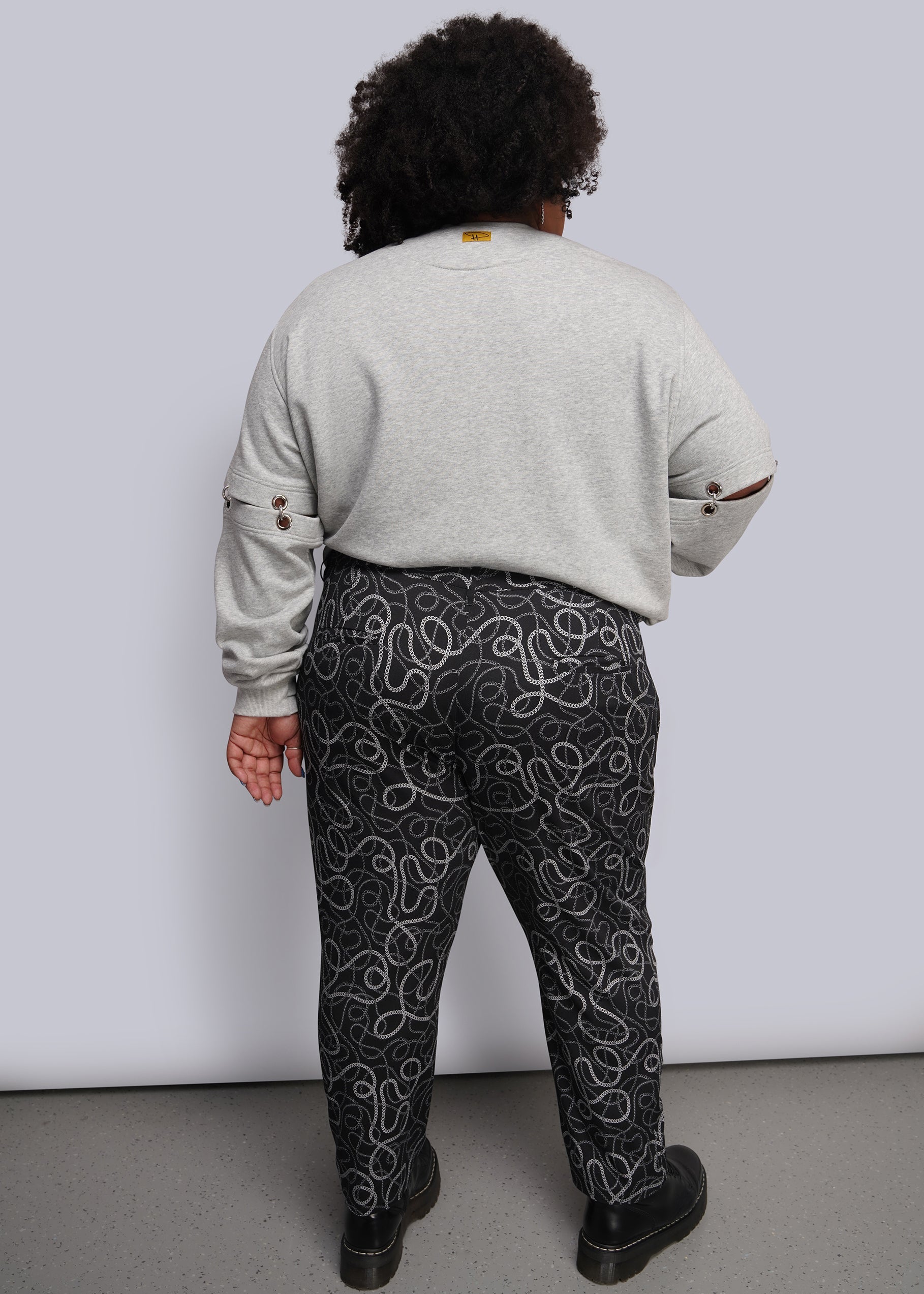 1X=A person shown from the back wearing a light gray sweatshirt and black pants with a white chain-link pattern, standing against a plain light gray wall.