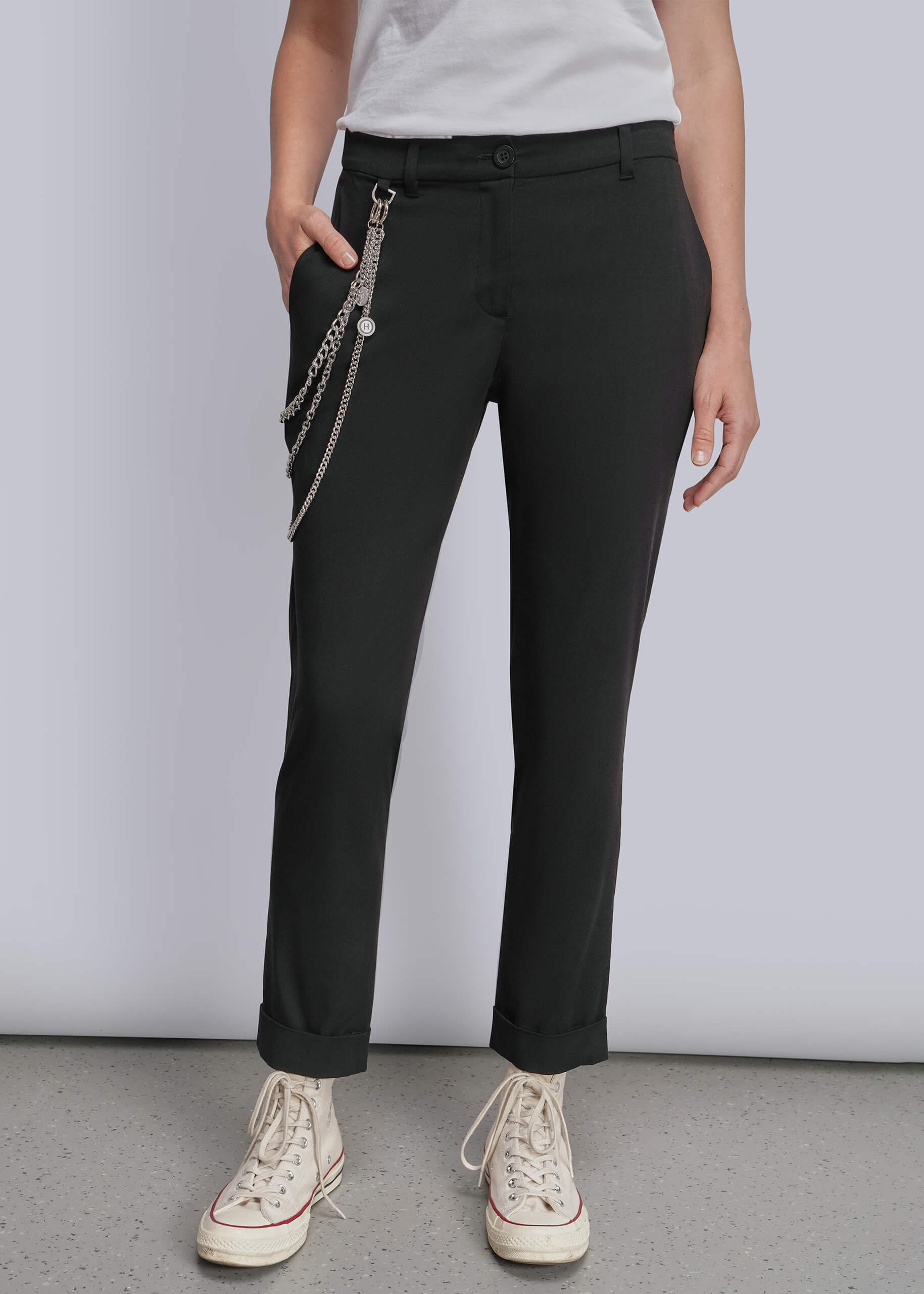 A model wearing black slim-fit trousers with a silver chain accessory hanging from the side pocket, styled with a white t-shirt and cream-colored canvas sneakers against a light gray studio background.