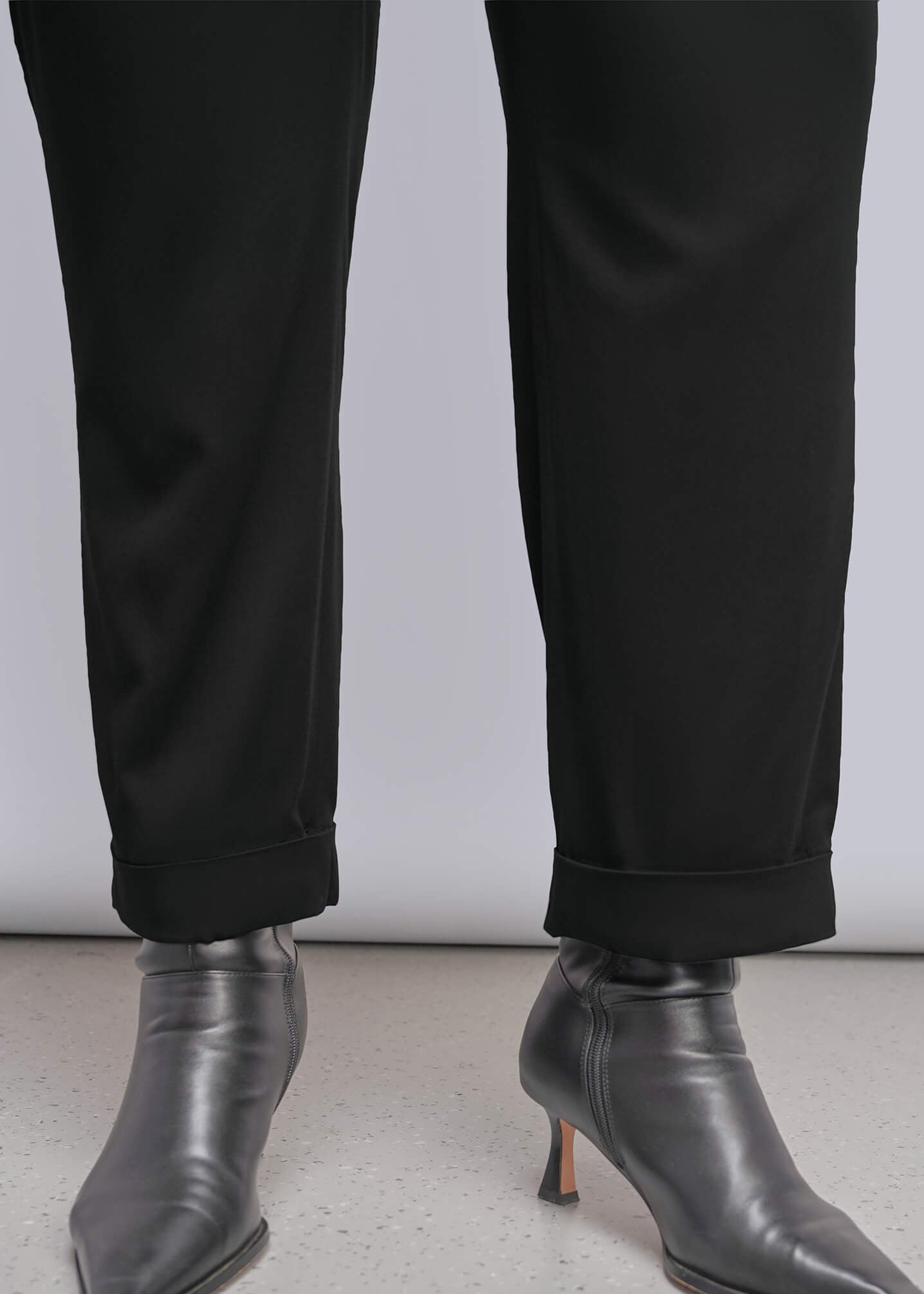 1X= A model wears black trousers that are cuffed and black leather pointed-toe ankle boots, shown against a light gray studio background.