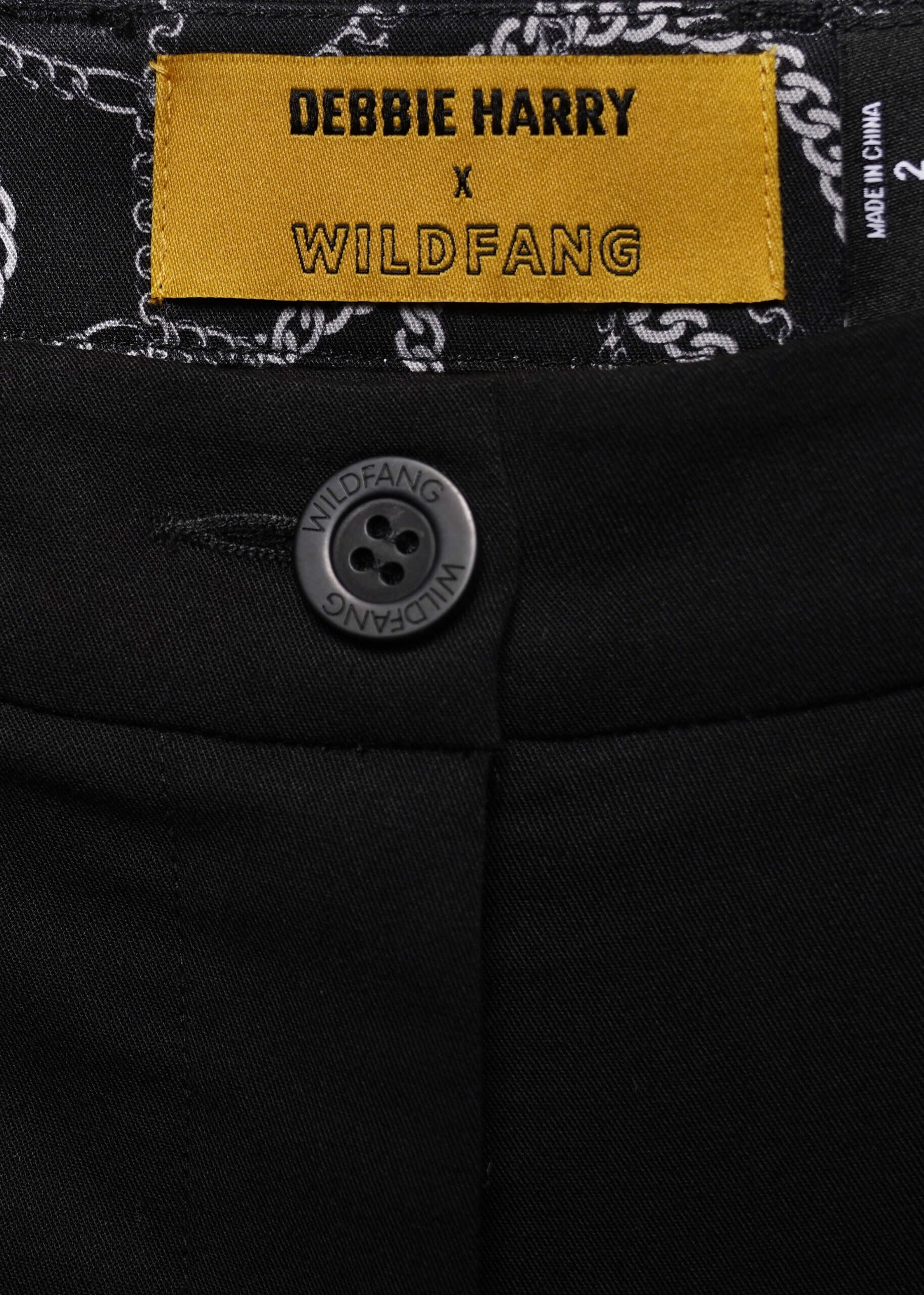 Close-up of black pants showing a yellow Debbie Harry x Wildfang brand label at the waistband and a round black button.