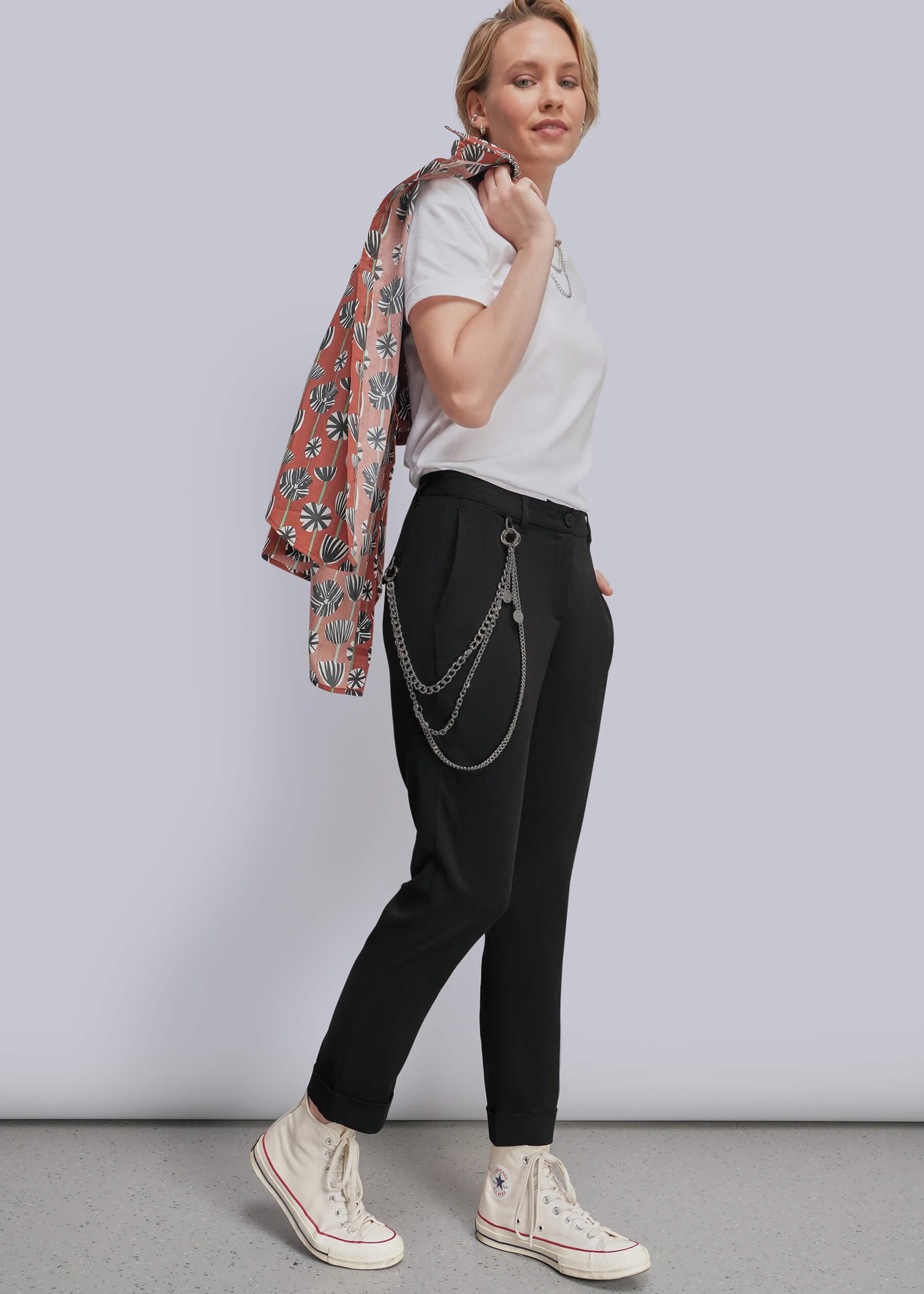 S=A woman modeling black trousers with a silver wallet chain accessory hanging from the waist, styled with a white t-shirt, cream sneakers, and holding a patterned button up over shoulder, against a light gray studio background.