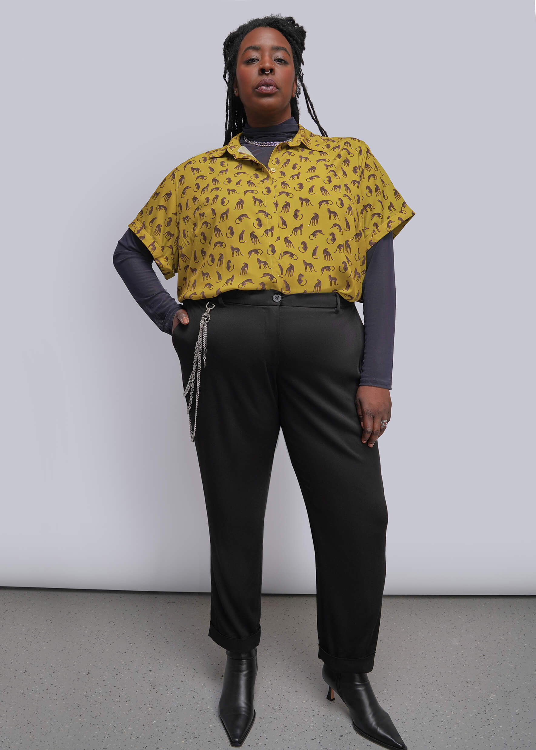 1X=A model stands against a light gray studio wall wearing a yellow short-sleeve button-up shirt with a small dark repeating pattern, layered over a dark gray long-sleeve top, black trousers, and black pointed ankle boots.