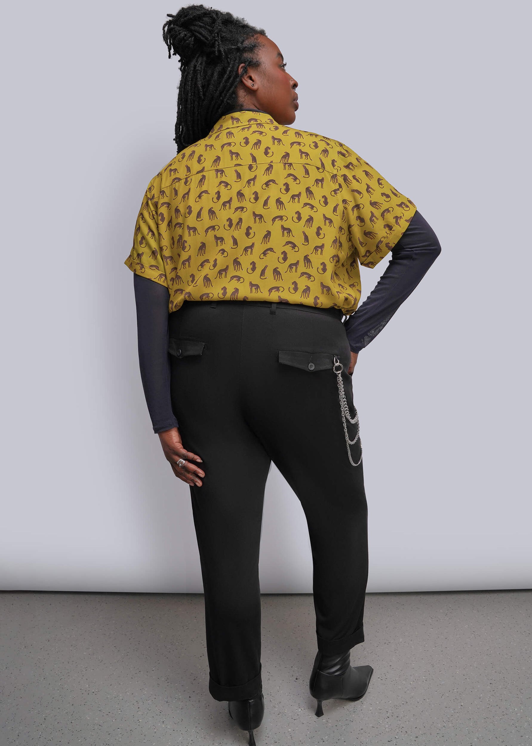 S=A model shown from the back wearing a yellow short-sleeve button-up shirt with a black abstract pattern, layered over a dark gray long-sleeve top, paired with black pants and black boots, against a light gray studio background.