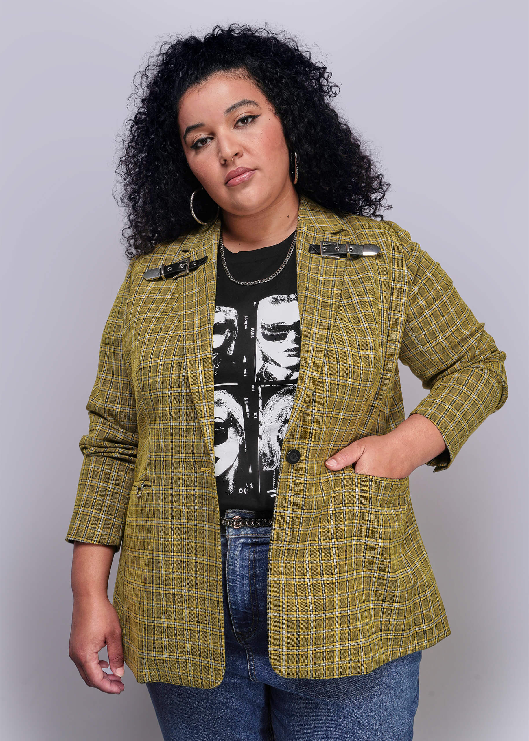 1X=Woman wearing a yellow plaid blazer over a black graphic t-shirt on a plain background