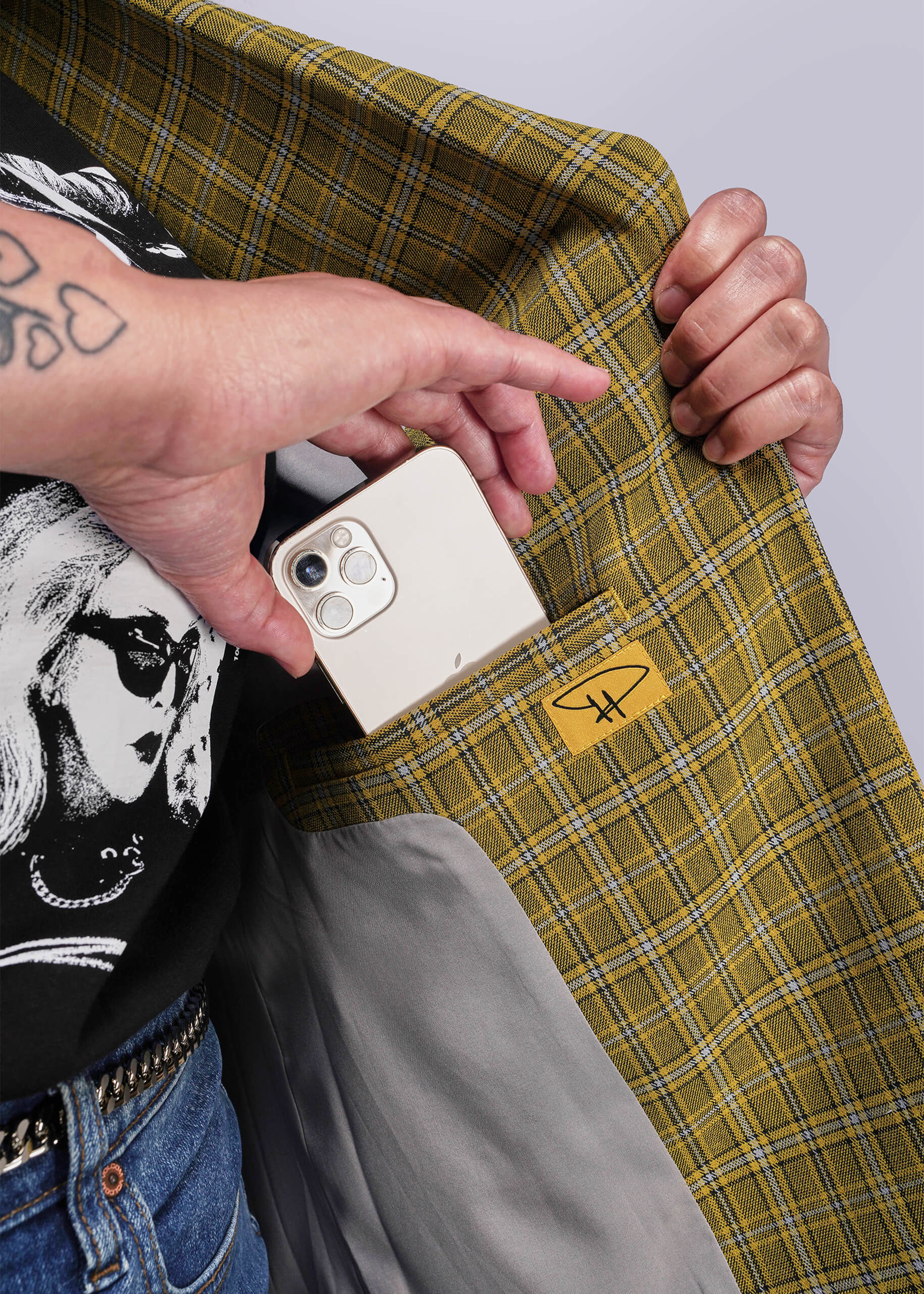 1X=Person holding a phone placing inside the inner pocket of a yellow plaid jacket with a Debbie Harry signature patch.