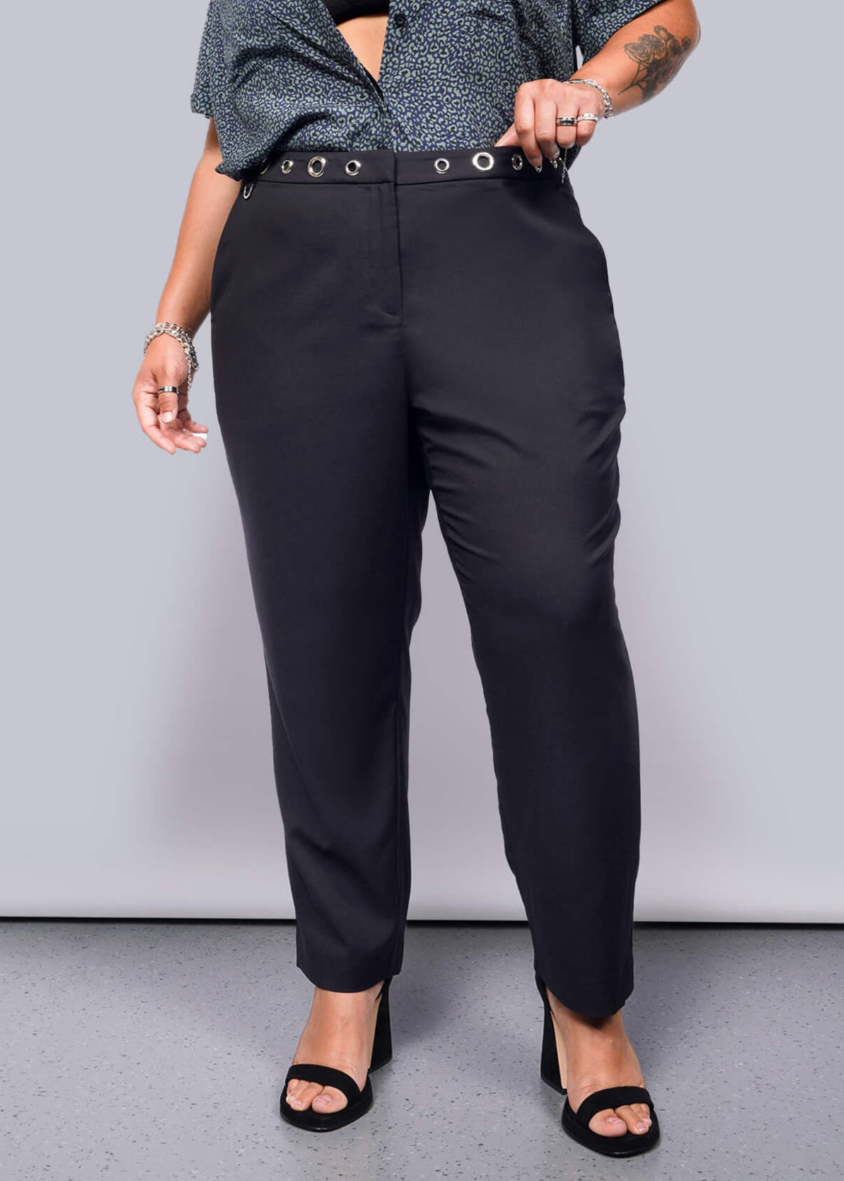 1X= A tattooed person on their left forearm wears Debbie Harry X Wildfang Slim Crop Pant in black, highlighted by large grommets, paired with a punk-style patterned blue shirt. They stand on a gray floor in black heeled sandals, with their right hand resting on the waistband.