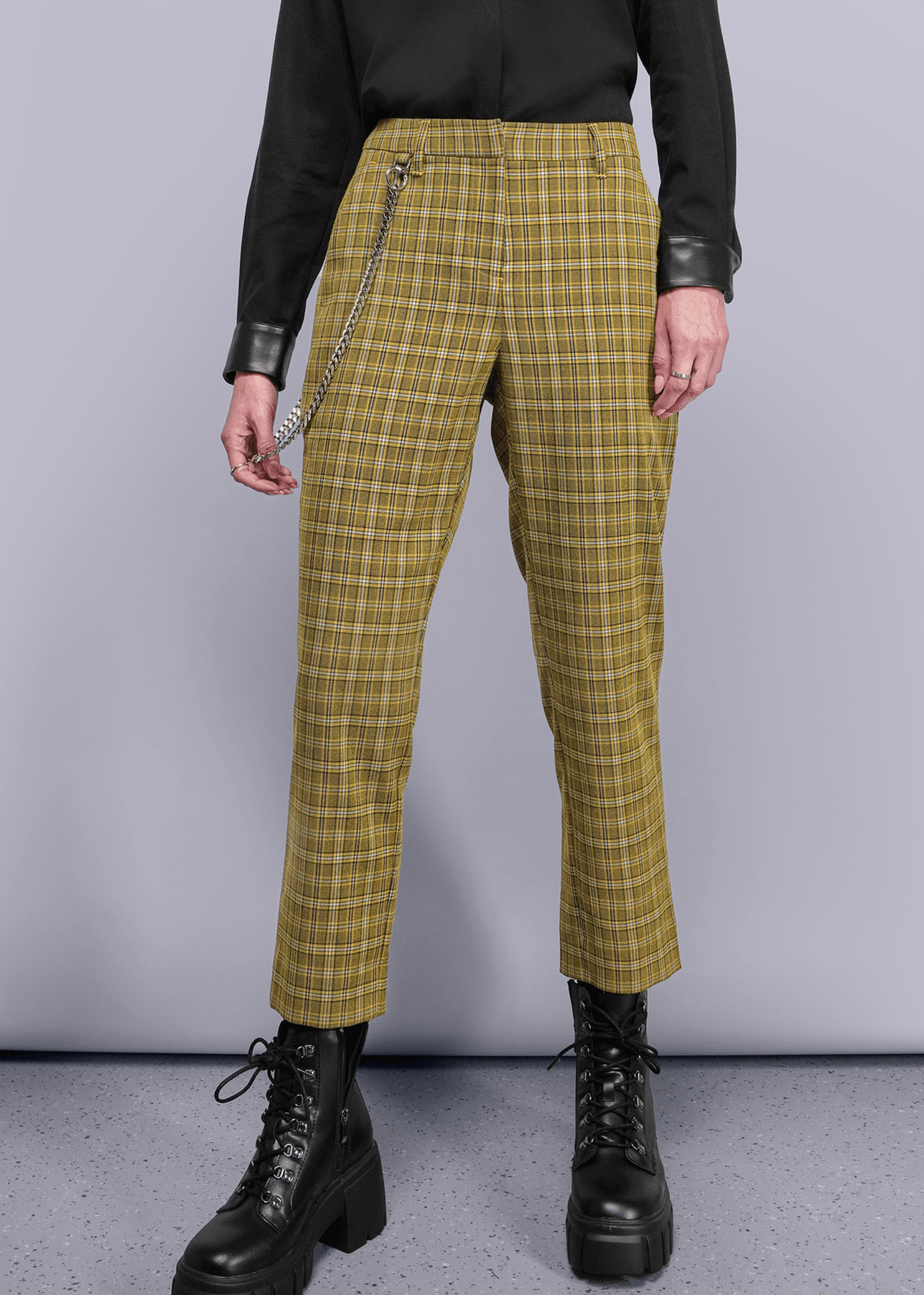 Person wearing yellow plaid pants with a black top and boots on a gray background