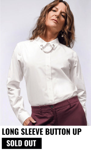 Model with brown wavy hair wears the white Debbie Harry x Wildfang long sleeve button up tucked into burgundy colored pants. Text underneath image reads LONG SLEEVE BUTTON UP and button reads SOLD OUT. White background.