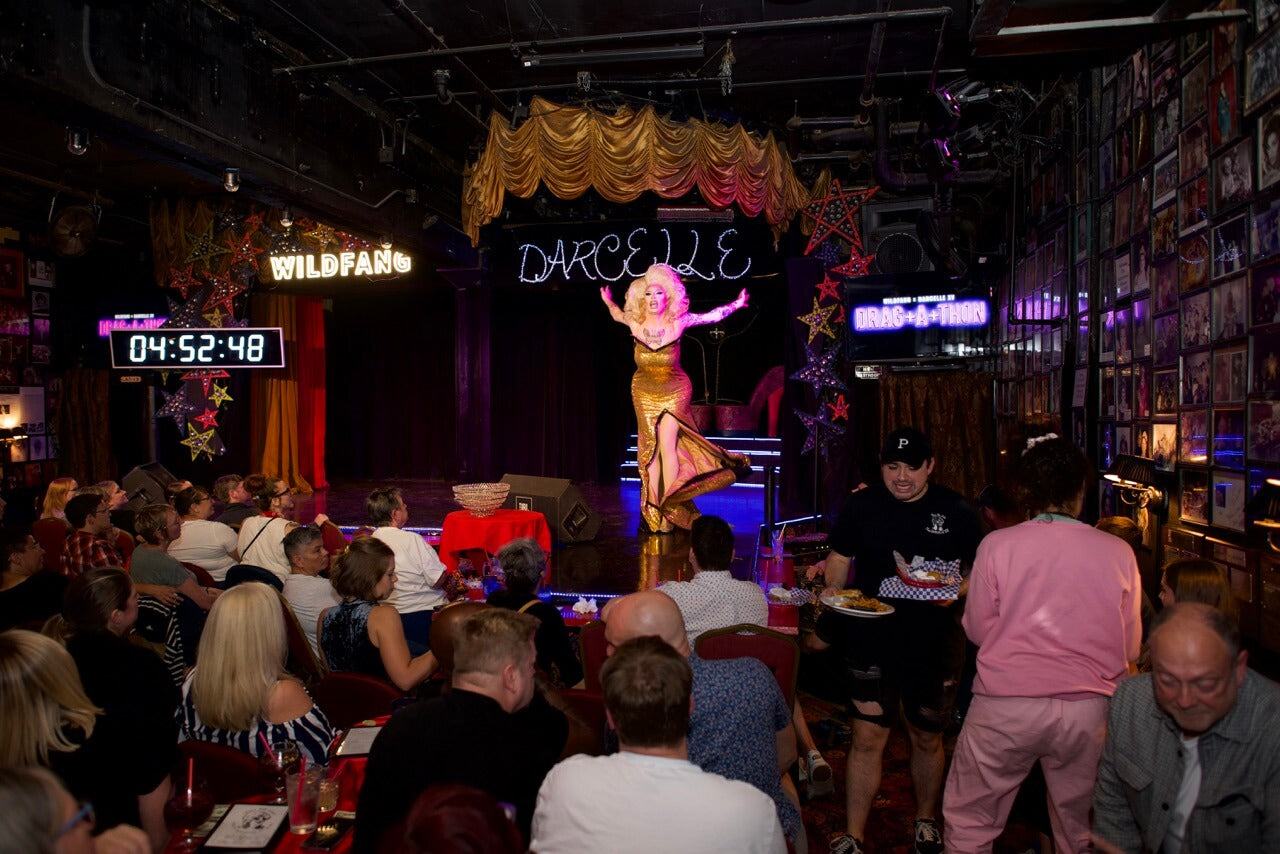 Drag queen in blonde wig and gold dress performs on stage at Darcelle's with crowd of seated people watching.